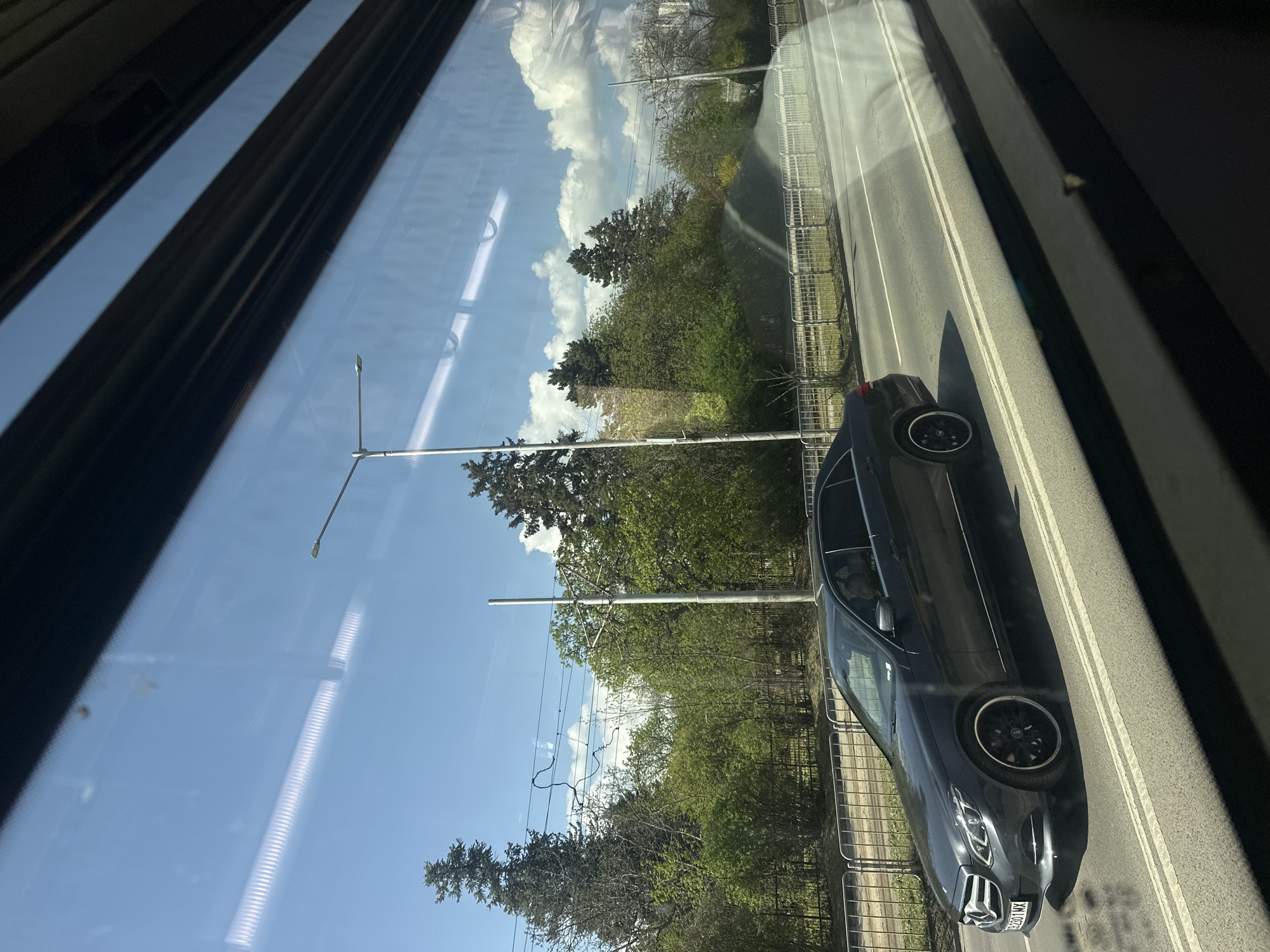 This image captures a vibrant street scene as viewed from inside a vehicle, likely a tram or bus, given the prominent window frame on the left and reflections of internal structures. The perspective is tilted, showing the exterior at an angle.
In the foreground and midground, a dark grey Mercedes-Benz sedan is visible on an asphalt road featuring clear double white lines. The car has black alloy wheels and a license plate reading "C 8201 MX," indicating it is registered in Sofia, Bulgaria. A person is faintly visible in the passenger seat of the Mercedes. Along the side of the road, a metal barrier fence separates the pavement from a lush green area filled with numerous trees and bushes, suggesting a park or green belt.
Overhead, tall utility poles with electrical wires crisscross the frame, characteristic of public transport infrastructure. The sky is bright blue with scattered, fluffy white cumulus clouds, indicating sunny weather. Strong sunlight casts clear shadows, suggesting it is mid-day or early afternoon. Reflections on the window glass obscure some details but also show the vehicle's interior elements, such as a dark patterned curtain or blind on the far left, and linear light fixtures from above within the transport.