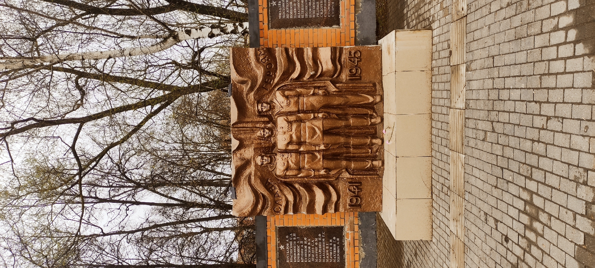 Here's a detailed description of the image:
**Overall Scene:**
The image shows a bronze war memorial in an outdoor setting. The memorial appears to be located in a public space, possibly a park or square, given the paved brick area and bare trees surrounding it.
**Memorial Details:**
* **Bronze Relief:** The central feature of the memorial is a bronze relief sculpture depicting three soldiers.
* **Inscription:** Flanking the bronze relief on either side are plaques with text inscribed on them. The text appears to be in Russian, and includes the names of people along with the years 1941 and 1945. This suggests the memorial is dedicated to those who served during World War II (specifically, the Eastern Front, also known as the Great Patriotic War in Russia).
* **Base:** The bronze relief rests on a light-colored stone base.
* **Paving:** The area immediately surrounding the memorial is paved with rectangular brick.
**Location (Inferred):**
Based on the visible details and the presence of the Russian text, the memorial is located in the city of Shchyolkovo, Russia.
**Environment:**
* **Trees:** Bare trees are seen in the background, indicating it is either late fall or early spring.
* **Weather:** The pavement appears wet, suggesting it has recently rained or is damp from morning dew.
**Additional Notes:**
* The memorial is likely a monument dedicated to soldiers who fought in World War II, representing an important historical event for the city and its people.
* There are a few pink flowers located at the base of the memorial. These are most likely artificial and meant to honor those that have fought in the war.