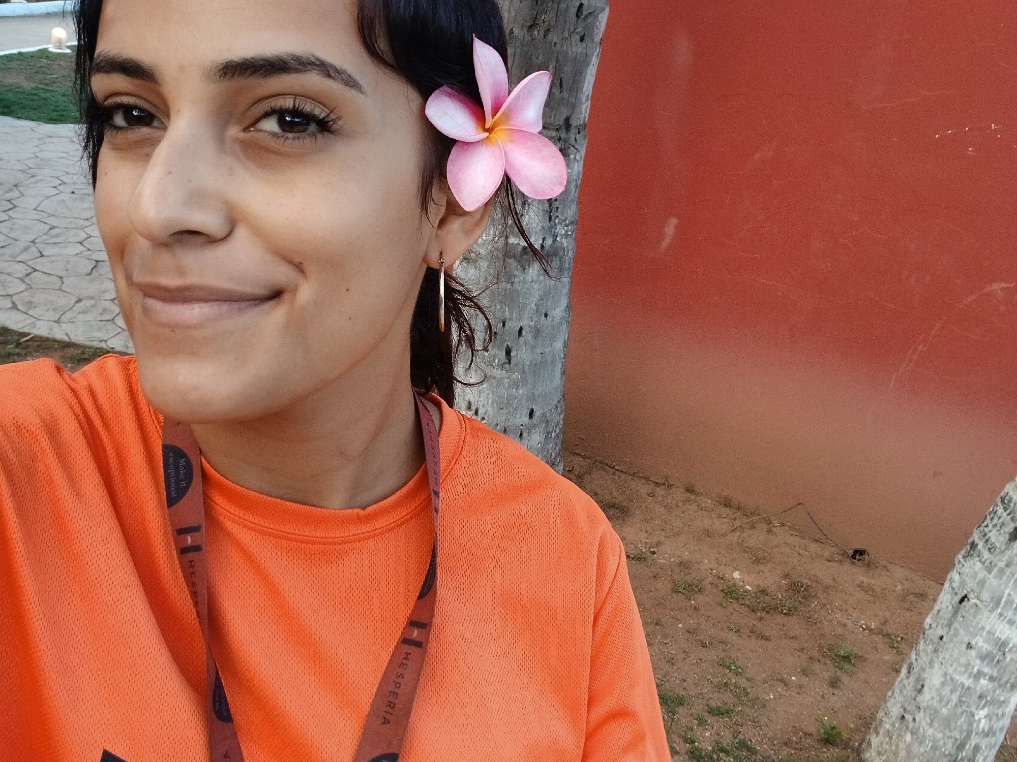 A close-up, slightly angled, selfie-style shot captures a young woman in Playa El Agua, Venezuela. She has dark hair, styled with a pink and yellow frangipani flower tucked behind her left ear and a thin hoop earring in the same ear. Her eyes are dark and appear to be looking directly at the camera. She has a slight smile. She is wearing a bright orange t-shirt with a lanyard from "Hesperia" hanging down the front. The background shows a grey, rough-barked tree trunk to her right, and behind that, a textured reddish-orange wall. To her left, a pathway with interlocking stone pavers and some sparse green grass is visible. The ground in the foreground appears to be dry dirt with scattered pebbles and small green plants.
