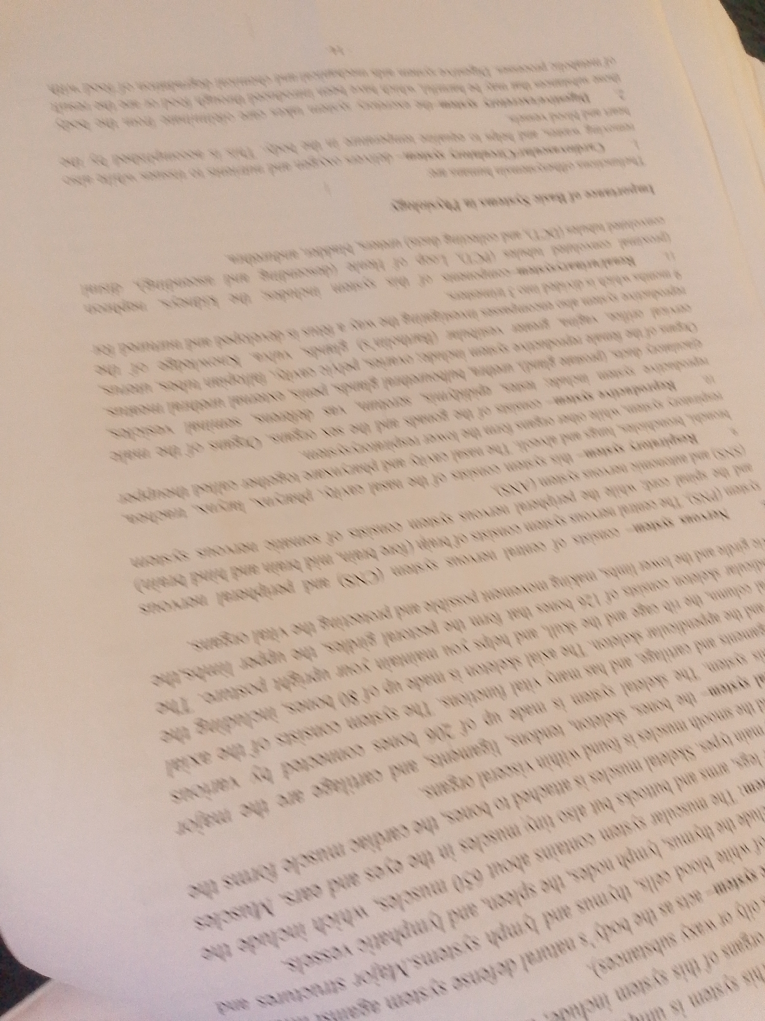 The image displays a close-up, angled view of a page from a textbook or study guide, focusing on a section detailing human anatomy and physiology. The text is printed in black ink on a light-colored paper, possibly white or off-white, and appears slightly faded or aged. The page is bookmarked, with a dark object, possibly a book or notebook, visible at the bottom right corner, casting a shadow and obscuring a portion of the page. The lighting suggests an indoor environment, likely a study area.
The visible text focuses on various body systems, including the nervous system, respiratory system, reproductive system, and skeletal system. It lists the components and functions of these systems. For example, the nervous system is described as consisting of the central nervous system (CNS) and peripheral nervous system (PNS), with the CNS comprising the brain and spinal cord. The skeletal system is noted to be made up of 206 bones. The section titled "Importance of Basic Systems in Physiology" is also partially visible. The text is dense, indicating a detailed explanation of biological concepts. There are no people, animals, or clear scene elements beyond the book page itself. The context is purely educational.