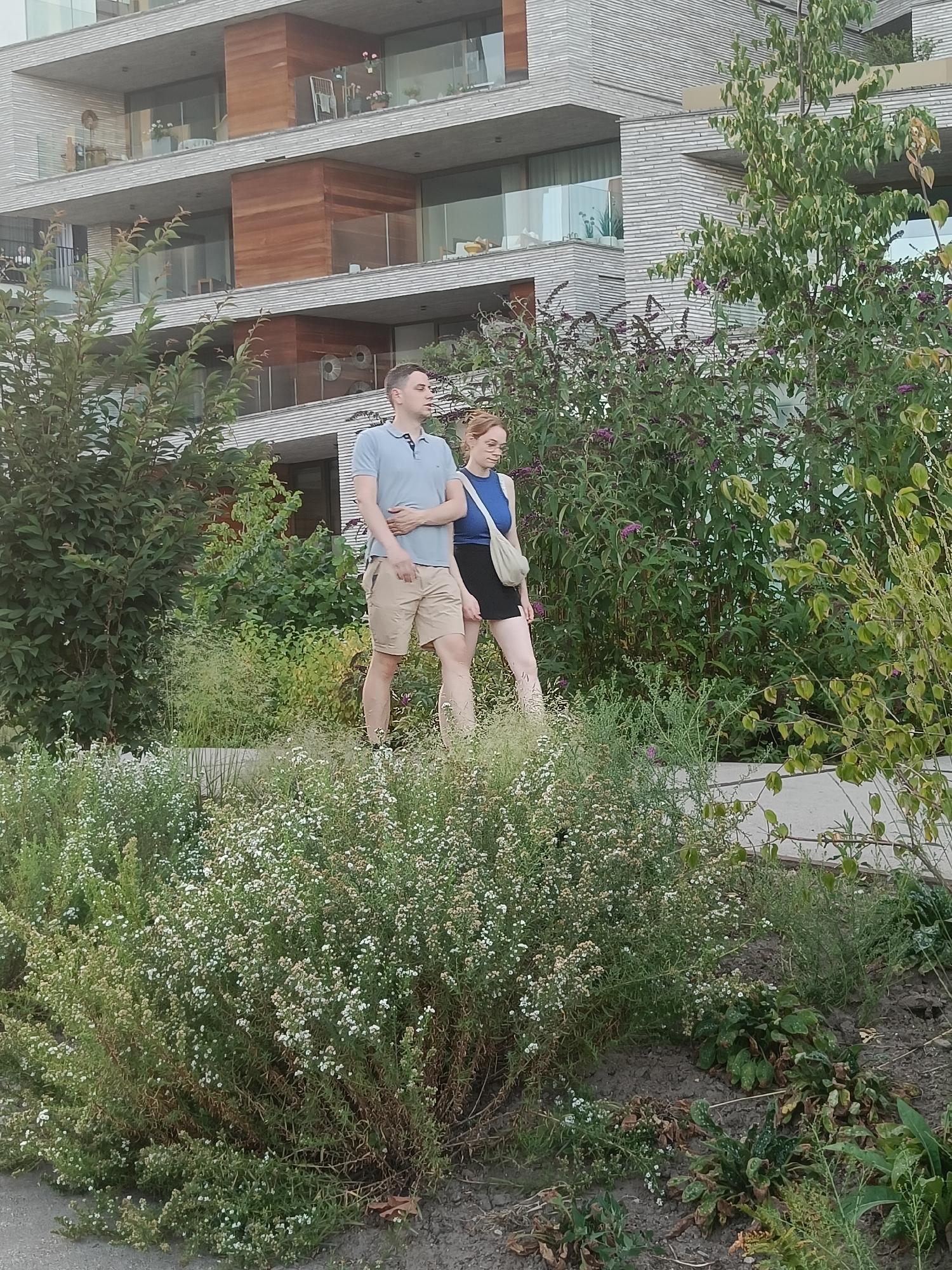 An outdoor scene depicts two people, a man and a woman, walking past a contemporary multi-story building with a facade of light-colored horizontal brickwork and wooden accents. The man, on the left, is wearing a light blue polo shirt and beige shorts. The woman, on the right, is wearing a dark blue sleeveless top and a black skirt, carrying a light-colored crossbody bag. They are walking on a paved path, with abundant green foliage and flowering bushes on either side. The building appears to be an apartment complex, with balconies visible on its upper floors. The greenery in the foreground consists of a mix of small white-flowering plants and taller, darker green shrubs. The ground is covered in soil and mulch. The lighting suggests it is daytime, likely late afternoon or early evening, given the soft light and shadows. The architectural style of the building and the landscaping suggest a modern urban or suburban setting. No specific landmarks confirm a location in Leuven, Belgium.