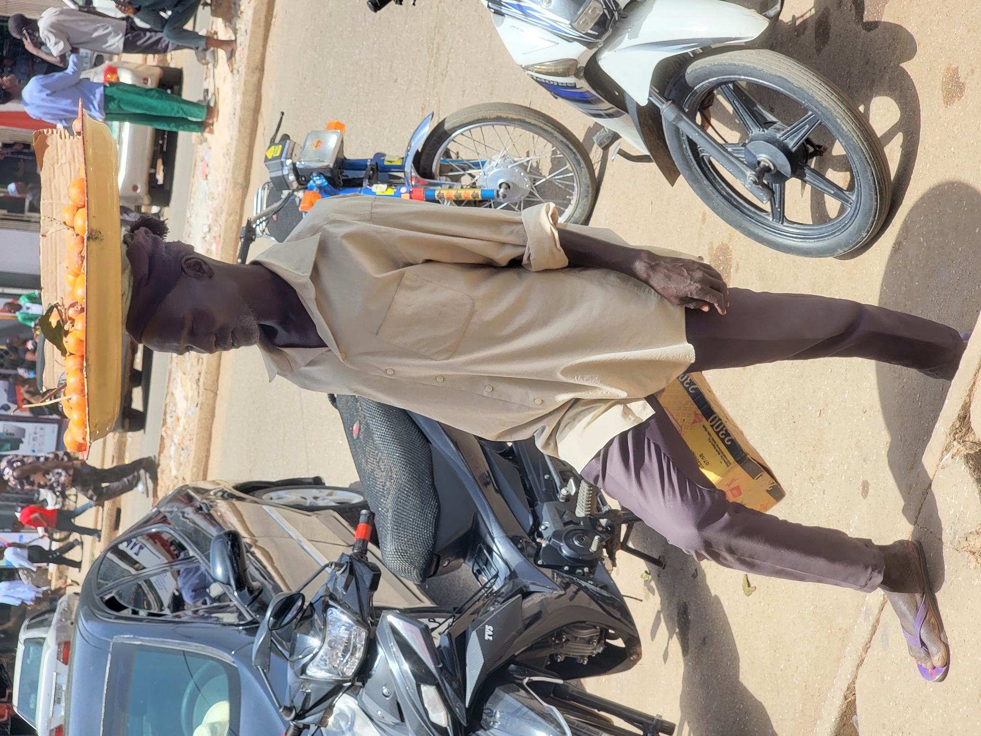 The image captures a street scene in Jos, Nigeria. A man is balancing a tray of oranges on his head while standing on the sidewalk. He is wearing a tan shirt, dark pants, and sandals. Beside him are parked motorcycles and cars. The scene suggests a typical day of street vending in Jos.