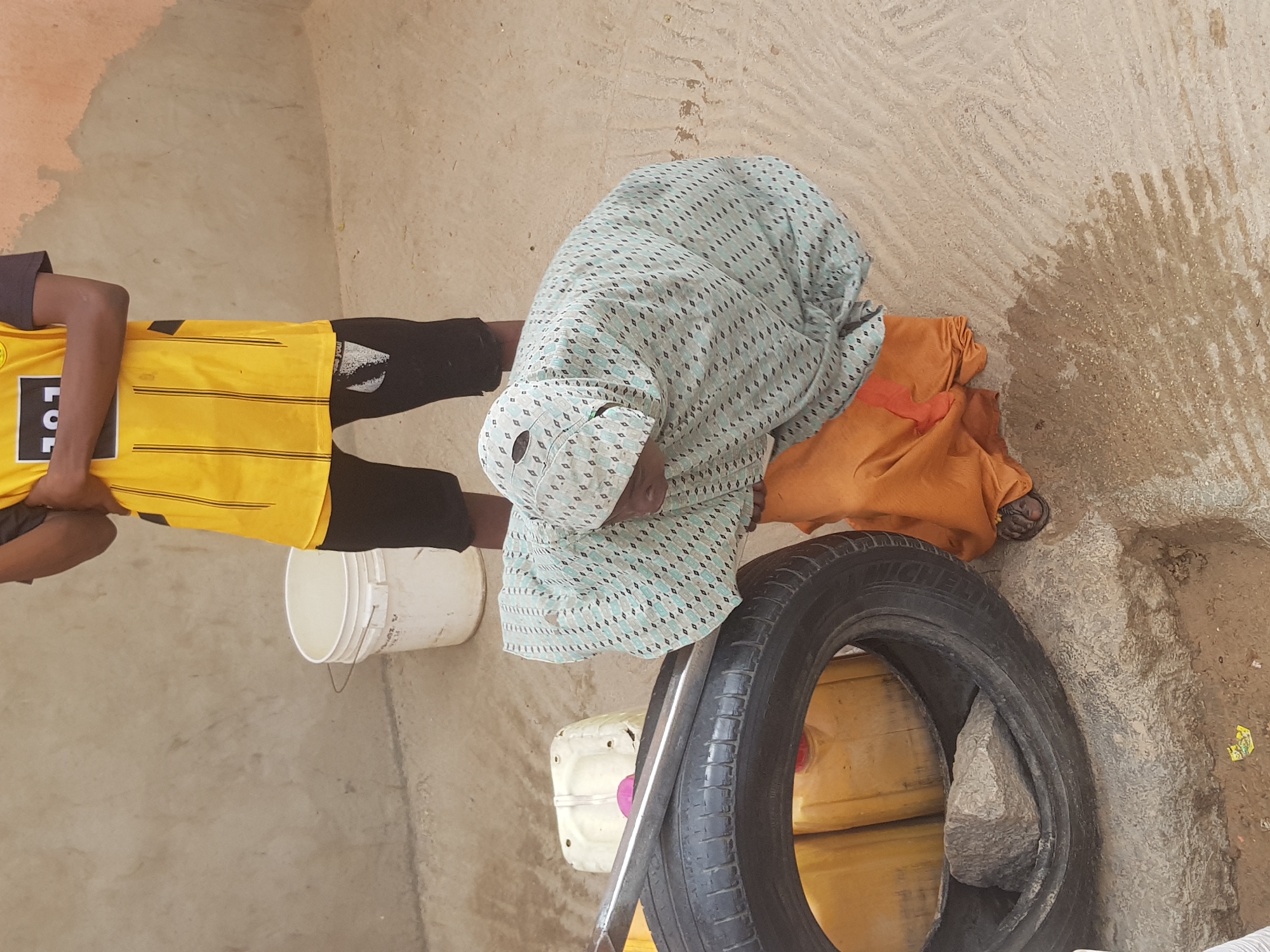 A person in a yellow and black shirt stands beside a white bucket, while another person in a patterned green head covering and orange fabric kneels near a black tire and yellow containers on a tan-colored surface in Dutsinma B, Nigeria.
