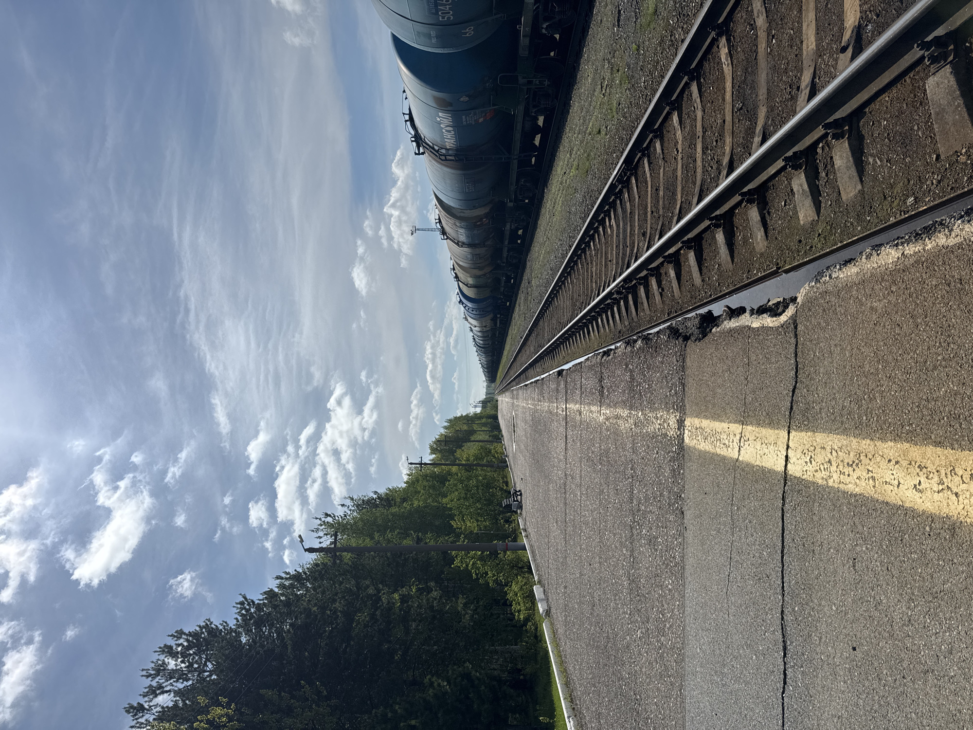 This media file captures a railway scene with a stationary freight train under a partly cloudy sky. The image is taken from a low angle, looking down the length of the railway tracks and a parallel platform.
The primary subject is a long freight train composed of numerous tank cars, predominantly blue or grey, stretching into the distance. Visible Cyrillic text on one of the tank cars clearly reads "ТРАНСОЙЛ" (TRANSOIL). Other partial numbers like "5046" and "5507" are visible on different cars. Two sets of railway tracks, featuring steel rails on concrete sleepers set in ballast, run parallel to the train.
In the foreground, an asphalt platform or paved area runs alongside the tracks, marked with a faded yellow line and showing cracks in its surface. To the left of the tracks, a dense line of green trees and bushes extends towards the horizon, indicating natural surroundings. Several utility poles with wires are interspersed among the trees.
The sky above is bright blue with scattered, wispy white clouds. The strong sunlight casts clear shadows, suggesting it is daytime, likely midday or early afternoon. The scene is static, with no evident activity or people. The overall impression is one of an industrial railway setting within a green landscape. The visible text "ТРАНСОЙЛ" aligns with the specified location context of Vysoky, Russia.