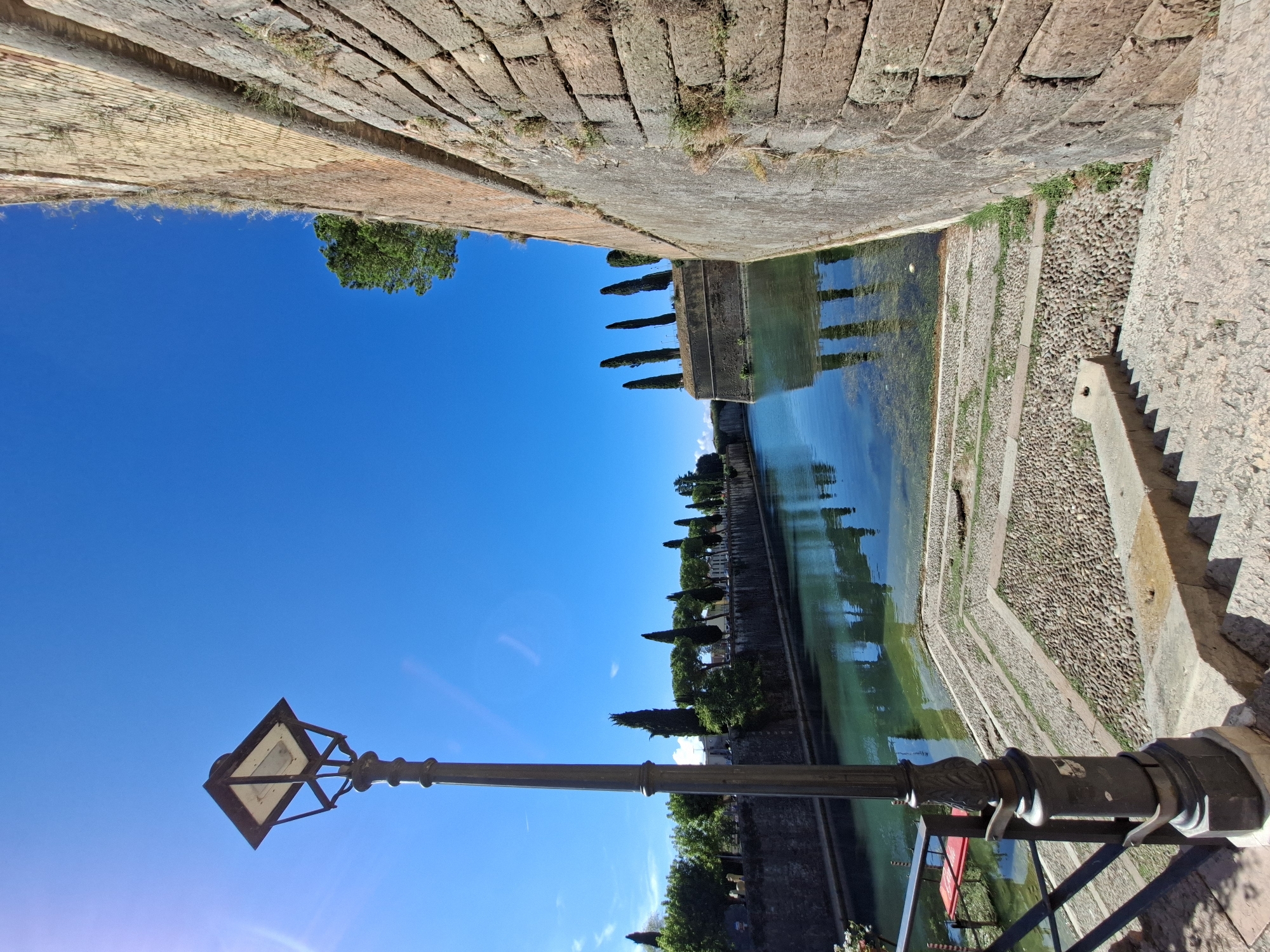 This is a full, eye-level, outdoor, wide-angle shot of a canal in Peschiera del Garda, Italy, with a strong, clear blue sky. In the foreground, a dark metal lamppost stands to the left of a set of stone stairs leading down to the water. The stairs are a light gray stone with some patches of cobblestone and grass between the risers. The canal, to the left of the stairs and lamppost, is a clear, turquoise color, reflecting the blue sky and surrounding greenery. Along the far bank of the canal, there is a stone wall topped with a row of trees, including tall, slender cypress trees. To the right of the canal, a massive, aged stone wall dominates the right side of the frame, composed of large, irregular blocks of yellowish-brown stone, with some grass and moss growing in the crevices. The sun is bright, casting a slight lens flare in the upper left quadrant of the image. There are no people or animals visible in the scene.