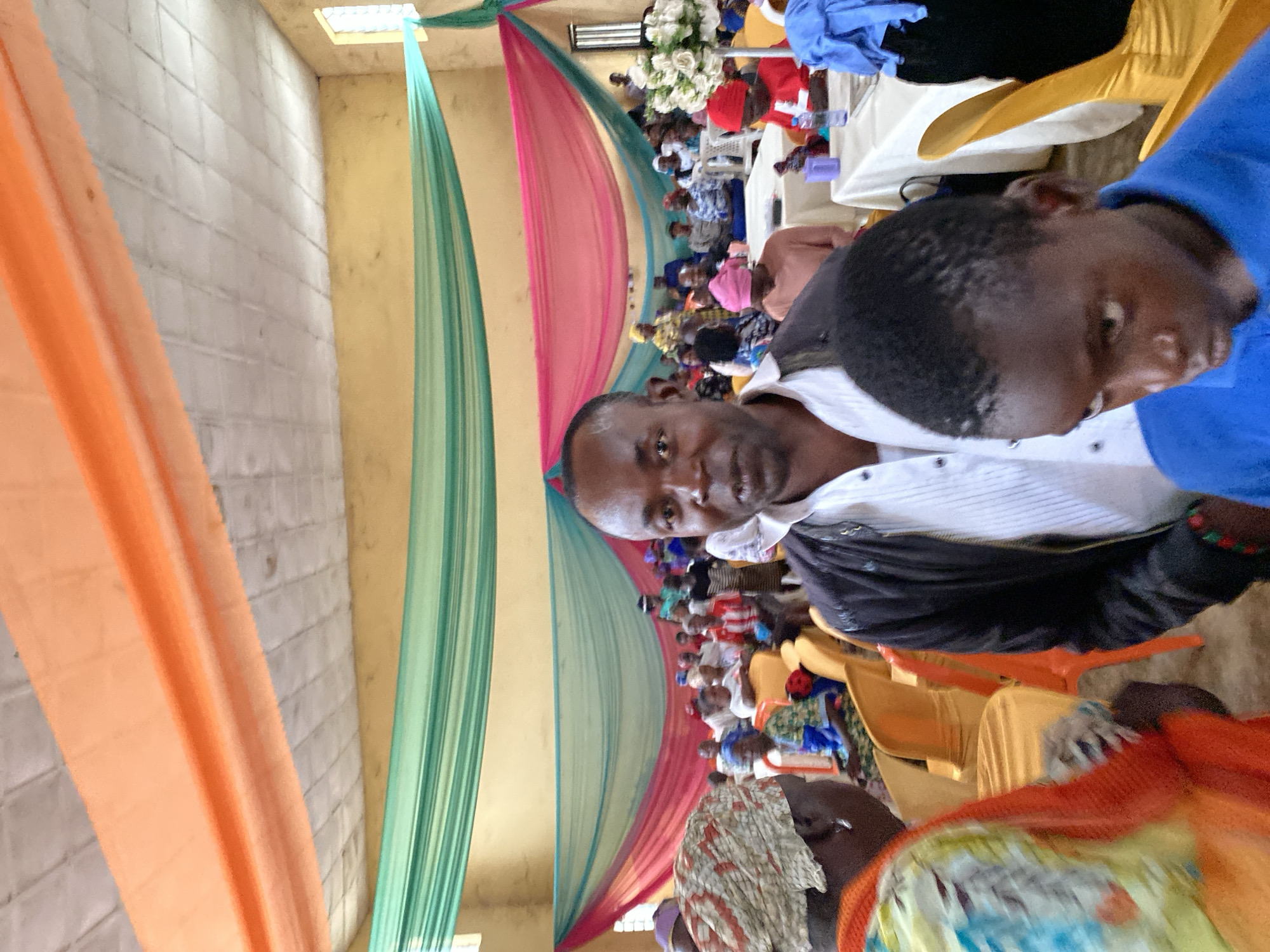 The image depicts an indoor gathering, likely a community event or service, taking place in a hall decorated with draped fabric in shades of orange, green, and pink. In the foreground, a man in a white shirt and black jacket is looking directly at the camera, with a younger person in a blue shirt partially visible in front of him. Behind them, a crowd of people, predominantly women, are seated in orange chairs, facing the front of the hall. Tables are set up with white tablecloths, and a floral arrangement is visible. The walls of the hall are painted a light yellow, and a window with barred panes is present. The ceiling appears to be a textured, concrete material. This scene is representative of communal events held in public spaces within Riyom, Nigeria.