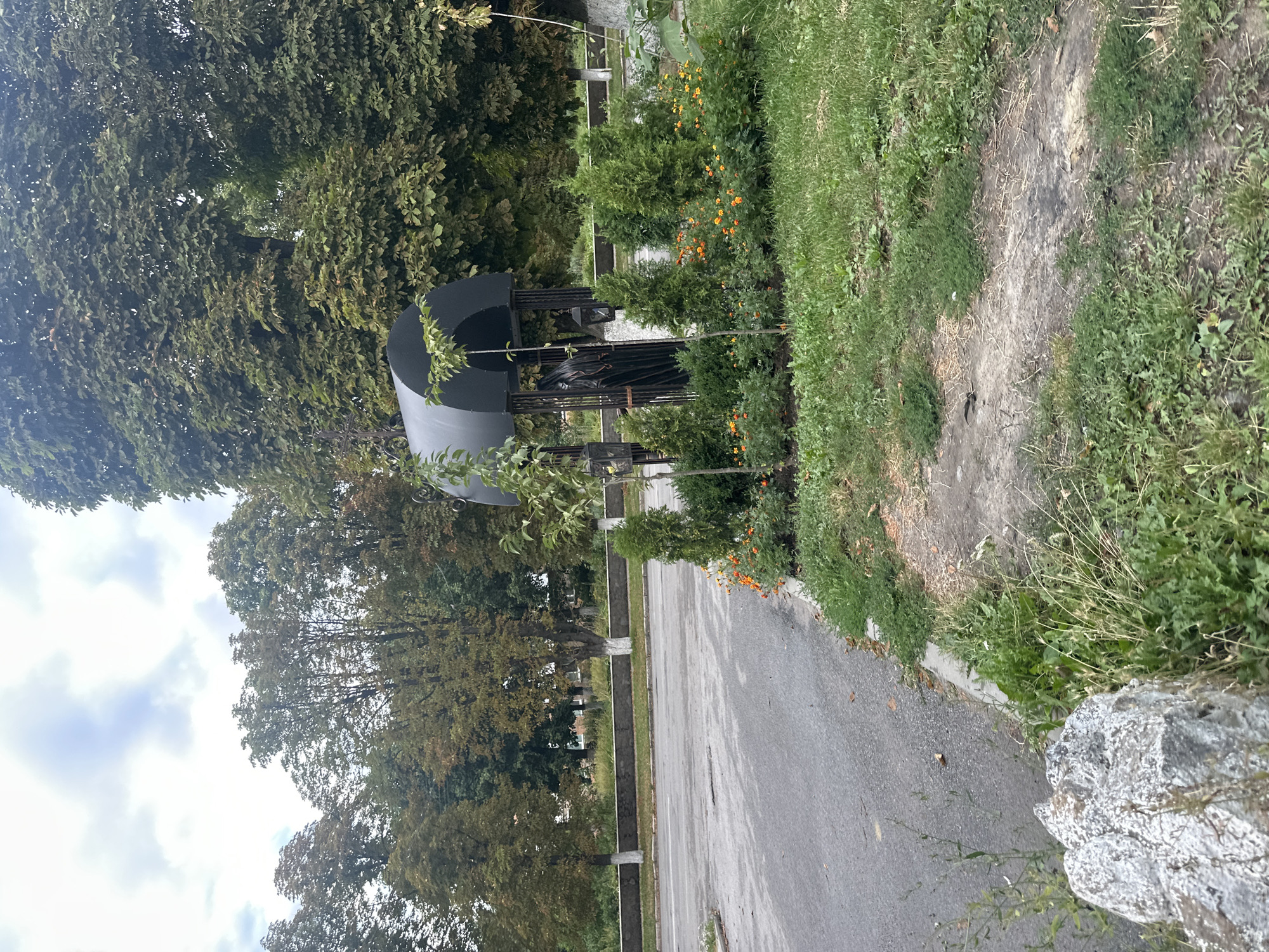 The image captures a street scene in Khmilnyk, Ukraine. In the foreground, a large gray rock sits on the left side of the frame, partially obscured by grass and weeds. A paved road occupies the lower left and center of the image, with a grassy shoulder bordering the right side. On the right side of the road, a well-maintained flower bed with orange marigolds and green shrubs is visible. Behind the flower bed is a black metal gazebo-like structure, enclosed with decorative metalwork. Inside the structure, a dark, draped figure, possibly a statue or effigy, is partially visible. Two hanging lanterns are attached to the exterior of the gazebo. Further back, a low stone wall with whitewashed tree trunks in front of it runs horizontally across the image. Beyond the wall, a line of trees with foliage in varying shades of green and brown forms a backdrop, suggesting it may be late summer or early autumn. The sky above is cloudy. The overall impression is one of a public space with decorative elements.