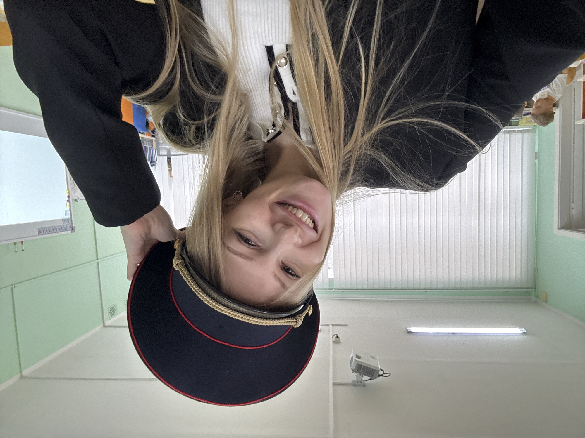 A young woman with blonde hair is holding a dark blue military-style hat with a red band and gold braid trim. She is smiling at the camera. She is wearing a black blazer over a white, buttoned-up top. The background appears to be an interior space with green walls and white blinds. A projector is mounted on the wall. The image was taken in a room with white ceiling and a florescent light. The location is the city of Perm, Russia.