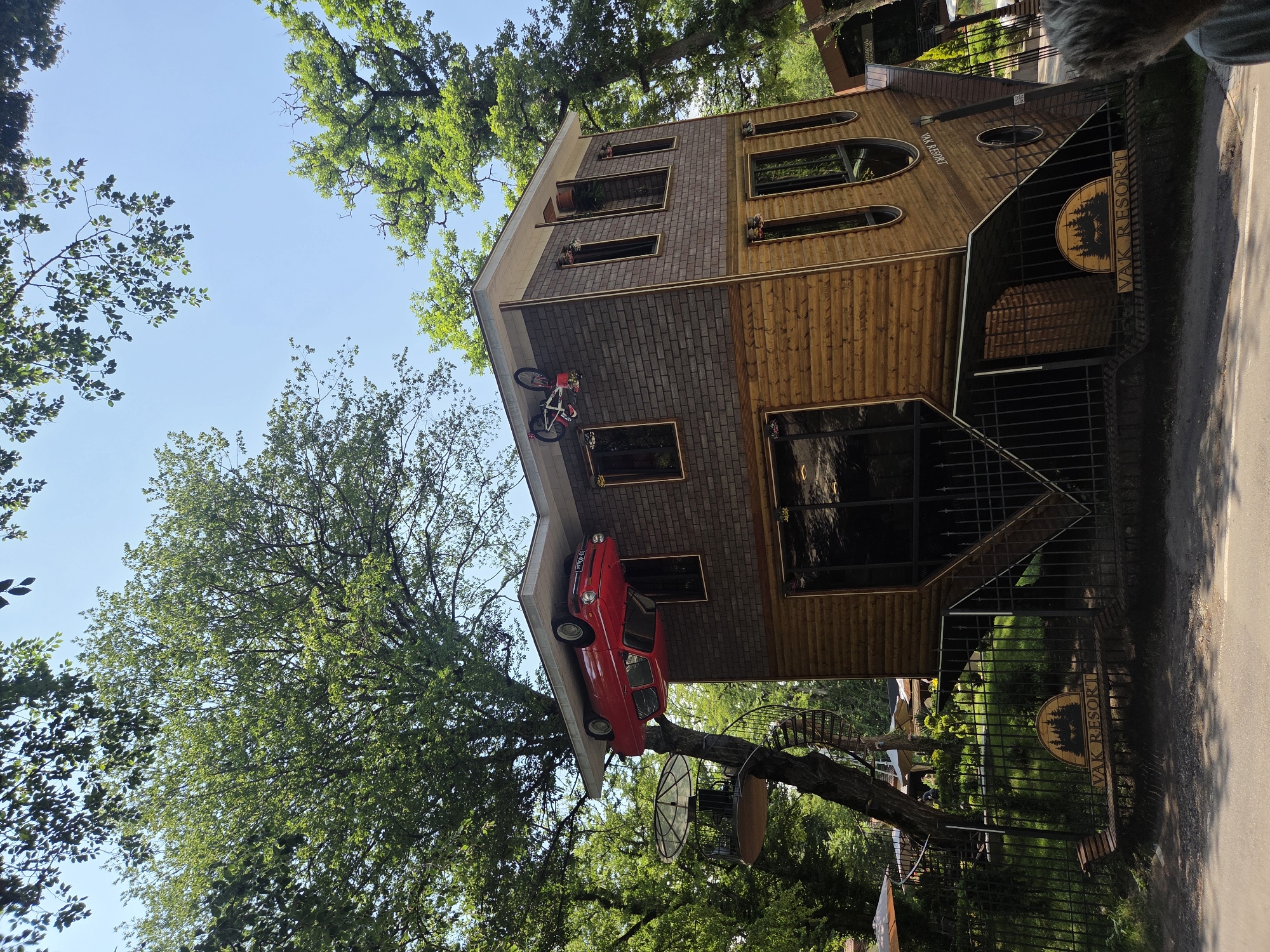 The image depicts an upside-down house, a unique architectural attraction located in Dilijan, Armenia. The house, designed to appear as if it's suspended in the air, features a red car on its roof and a bicycle on the side. The structure is surrounded by lush green trees and a clear blue sky, suggesting a natural and serene environment. The house has two stories, with the lower floor appearing to be the foundation. The exterior walls are a mix of wooden planks and brick-like material. A black metal fence encloses the property, and a bench with a sign reading "VAK RESORT" is visible in front of the fence. The setting appears to be a resort or tourist destination, given the presence of the sign and the overall aesthetic. The image is taken from a slightly low angle, emphasizing the unusual orientation of the house.