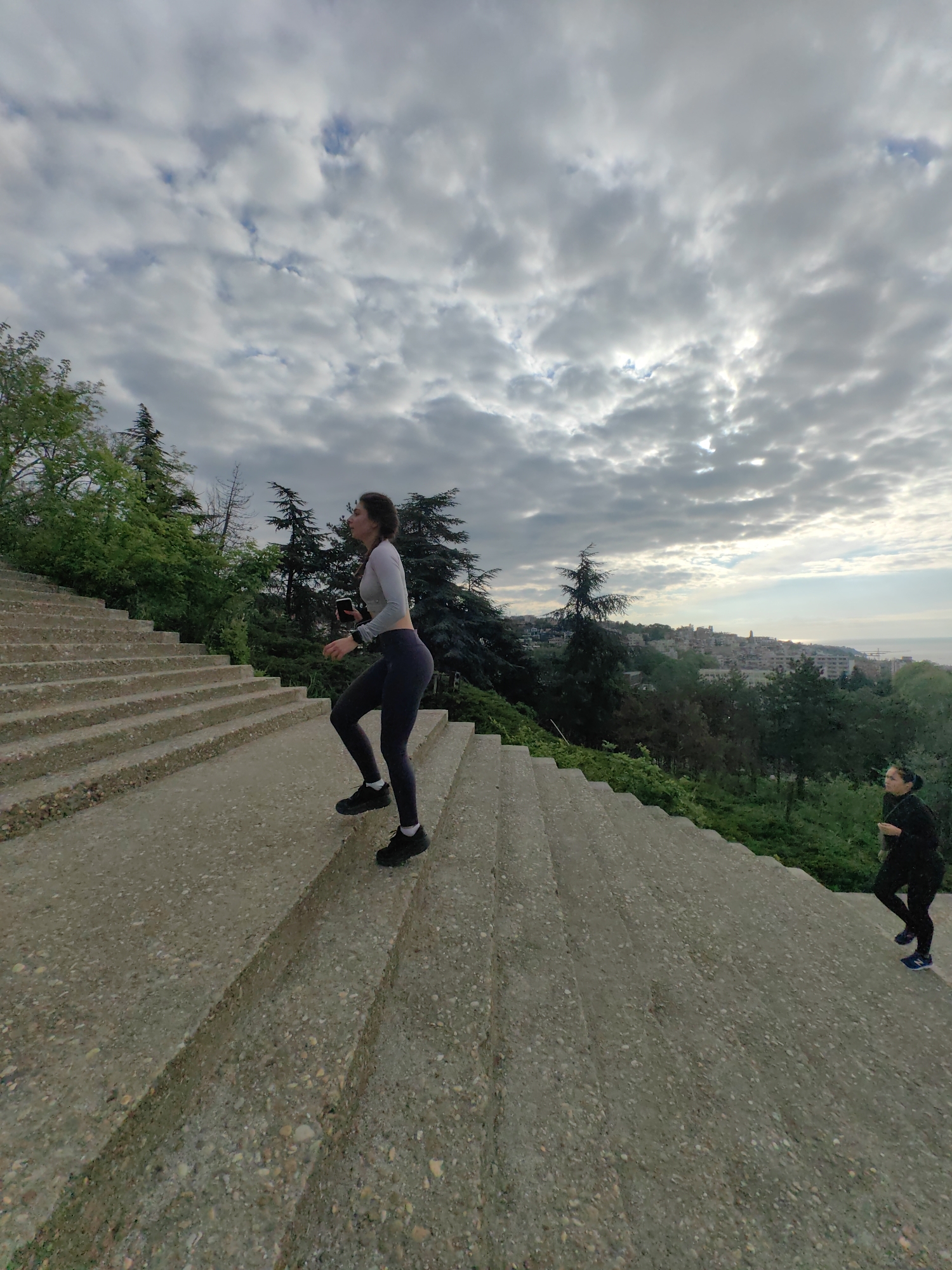 The image shows a young woman in athletic wear running up a long, concrete staircase in Varna, Bulgaria. She wears a light-colored long-sleeved top, dark leggings, and black sneakers. She holds a smartphone in her hand. Another person in dark athletic wear is running up the stairs further back. The steps ascend towards a view of the city and the Black Sea under a cloudy sky. Green trees line the sides of the staircase. The photo captures an outdoor fitness activity in an urban environment.