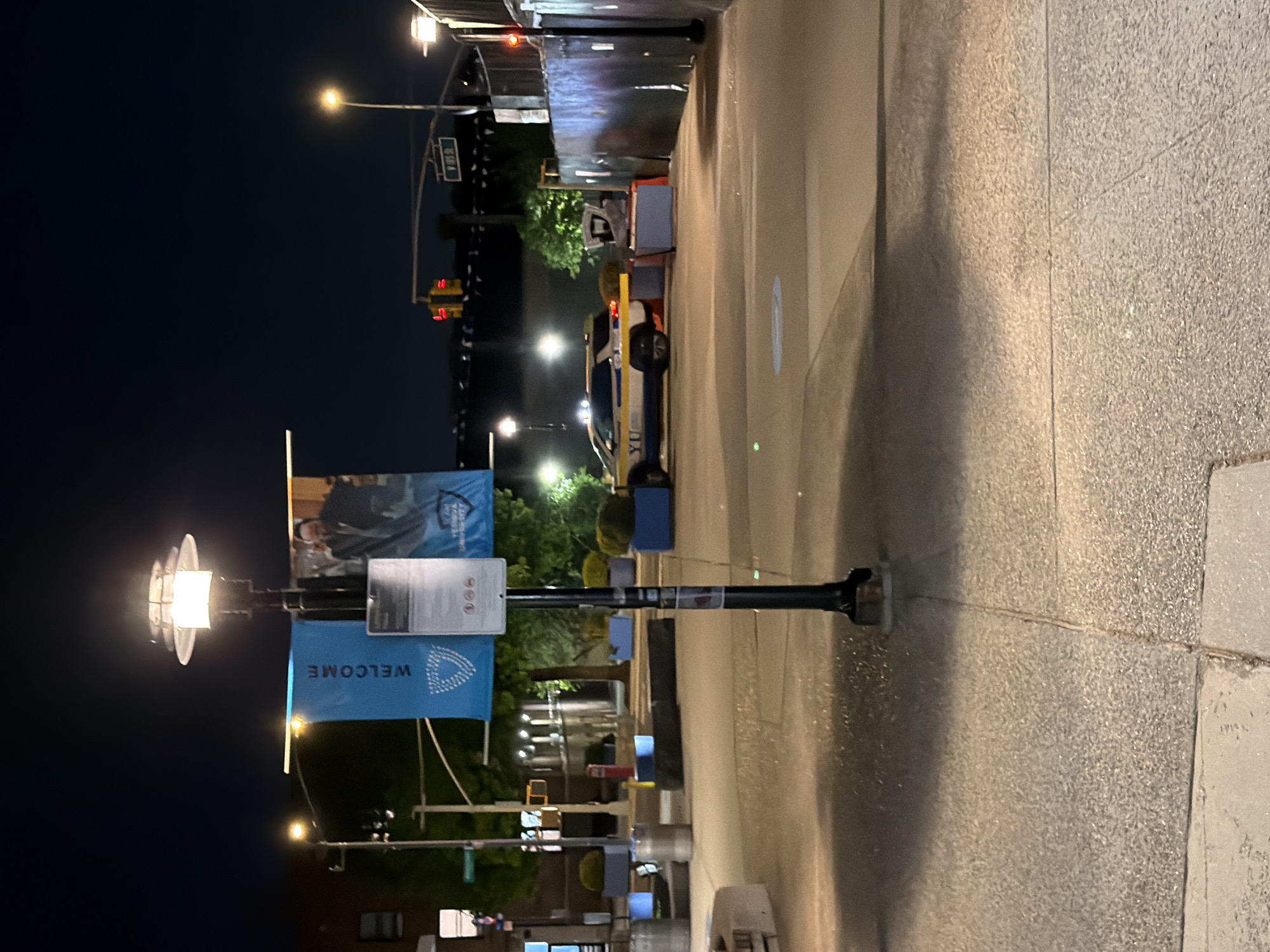 Here's a detailed description of the image:
**Location:** The image appears to be taken on a street at the Yeshiva University campus at night in New York City, United States of America.
**Scene:**
The scene shows a street view at night, illuminated by streetlights and artificial lighting.
A lamppost is prominent in the foreground. Banners are hanging off the lamppost; the word "WELCOME" is visible on one. Other advertisements or signage can be seen further up the pole.
A building is on the left side of the image.
A university security vehicle parked on the right.
On the left side in the background, a traffic light can be seen. There is a street sign showing the intersection with West 185th street.
The pavement of the street is visible, with a sidewalk on the right.
The sky is dark, suggesting nighttime.