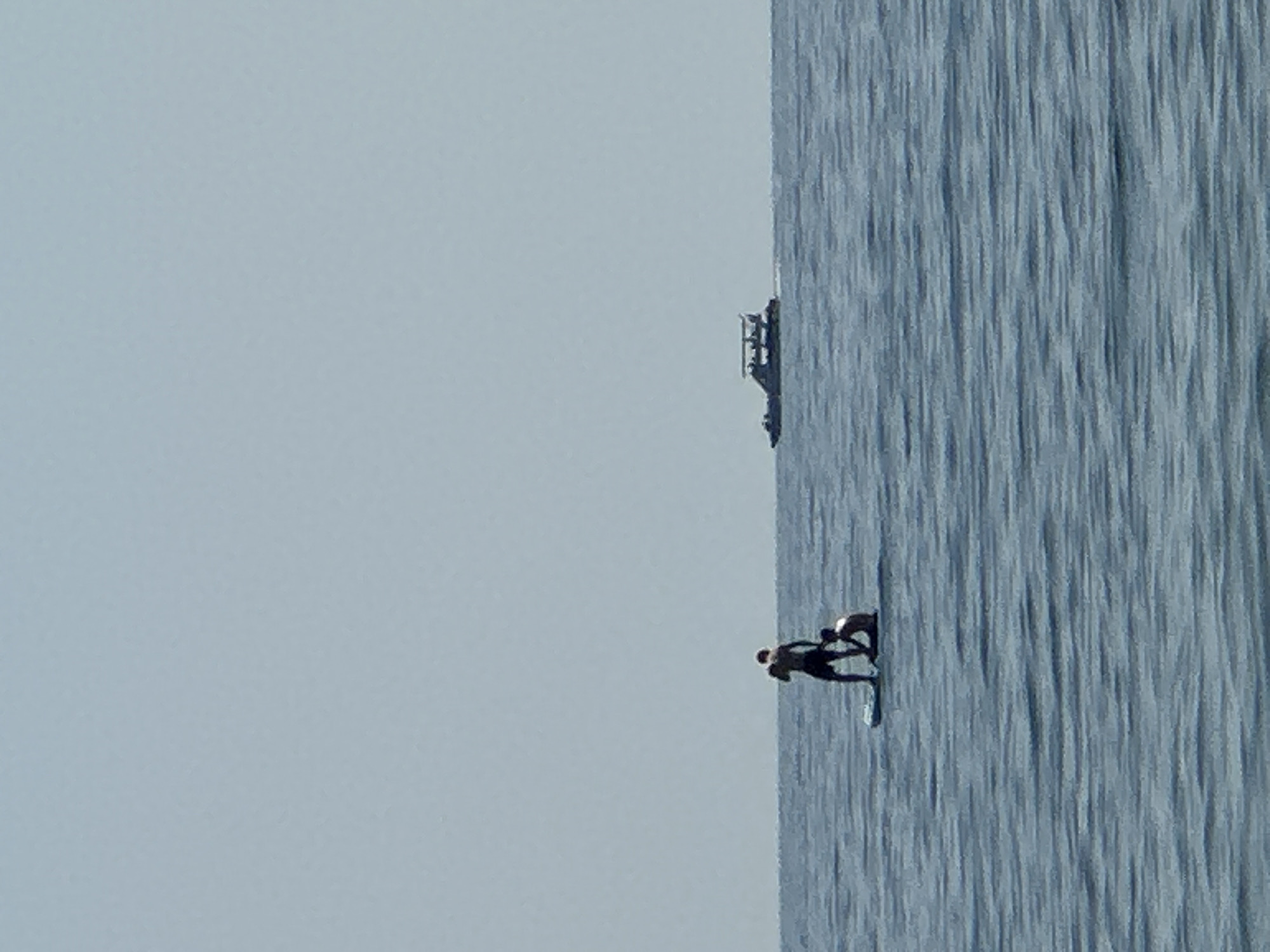 Two people are on a paddleboard in the sea near Đurovići, Montenegro. The person in front is standing and holding a paddle, while the person behind is kneeling. A boat is visible in the distance. The water is calm with small waves, and the sky is clear and pale blue. The scene appears to be during daylight.