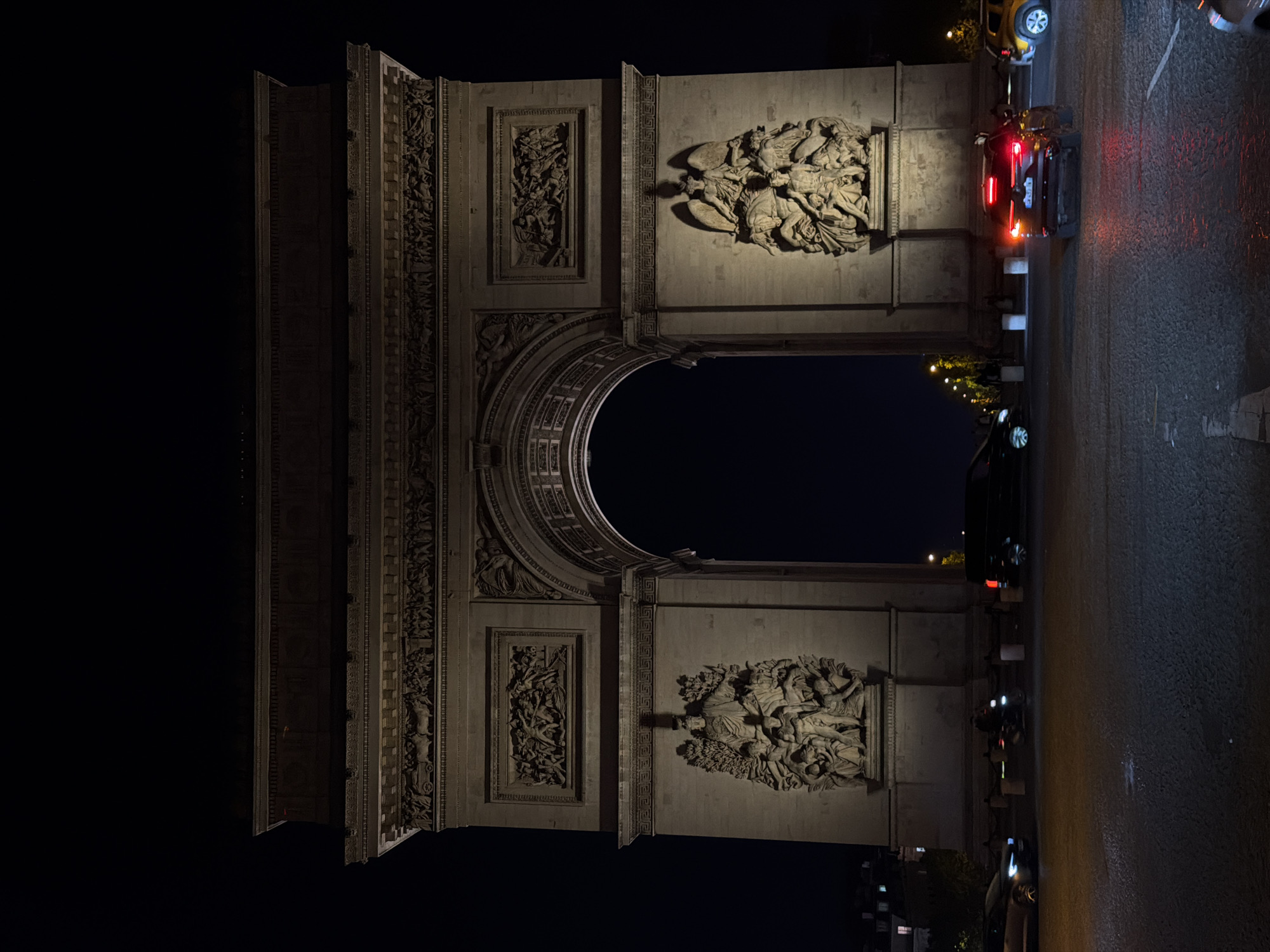 The Arc de Triomphe, a monument in Paris, France, is seen at night. The arch is illuminated. Detailed sculptures adorn the structure. A roadway runs along the right side of the image with vehicles driving by. The night sky is visible through the archway.