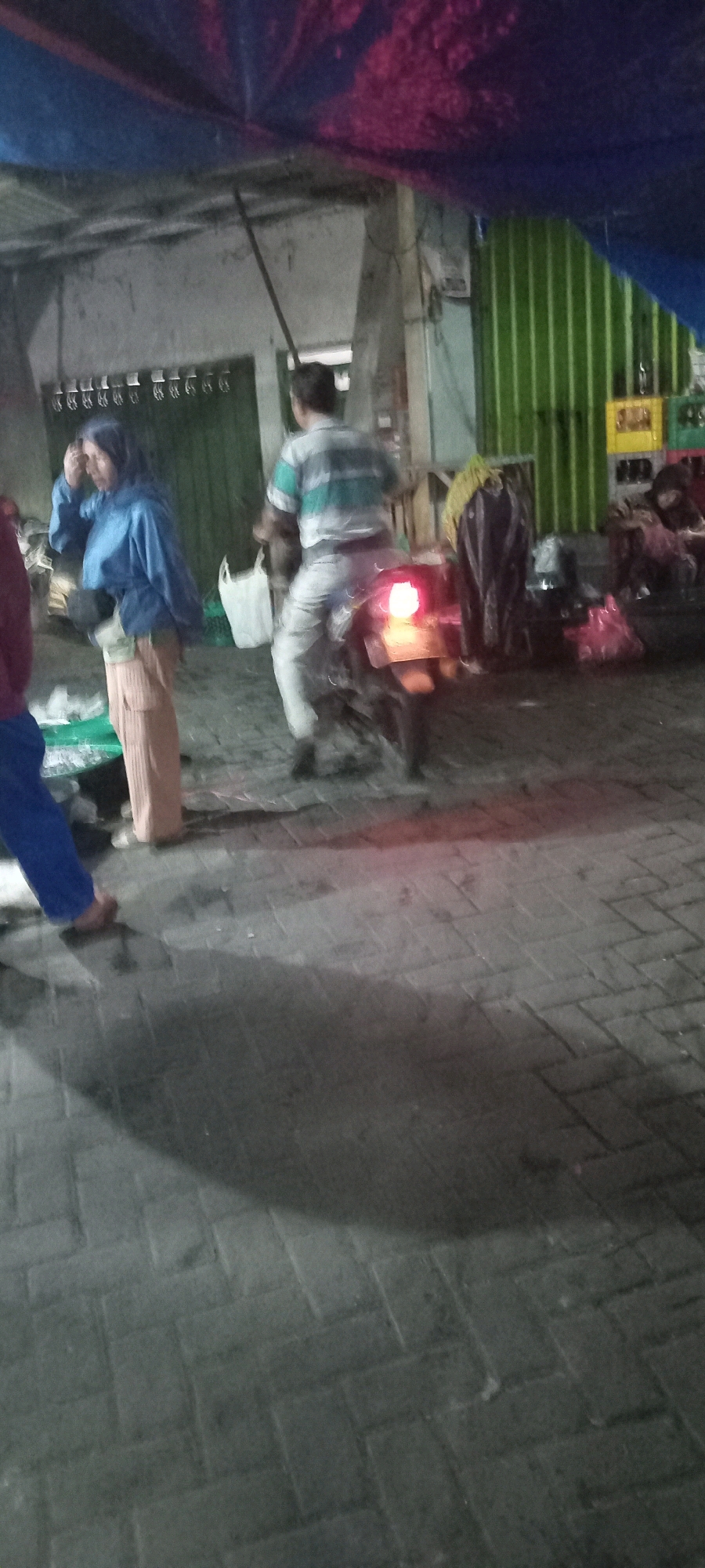 The image shows a street scene in Babat, Indonesia, at what appears to be dusk or nighttime. In the foreground, a person wearing blue pants and a light-colored shoe is partially visible on the left. To their right, a person in a blue jacket and light-colored pants stands, holding what looks like a black object and a white plastic bag. Behind them, a person on a motorcycle is facing away from the camera. The motorcycle has a lit taillight. The background features a green corrugated metal wall and various stalls or structures with tarps. The ground is paved with interlocking bricks. The overall impression is of a marketplace or street vendor area.