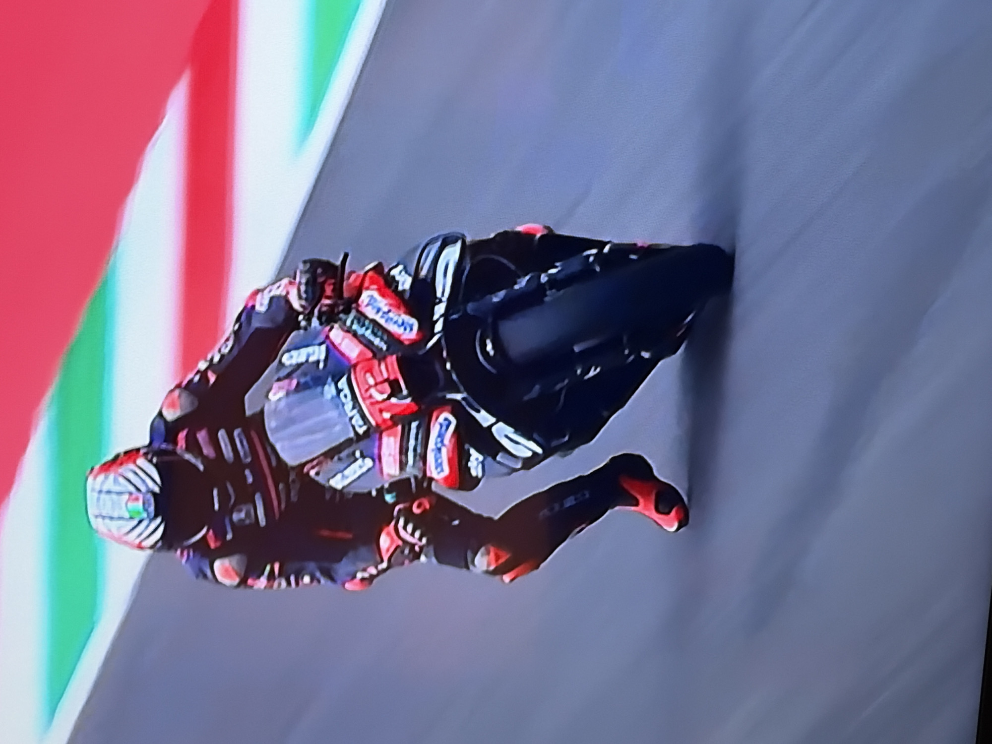 A motorcyclist is leaning sharply to the left while riding on a gray asphalt track, with the Italian flag (green, white, and red stripes) blurred in the background. The rider wears a black, red, and white racing suit and helmet, and is positioned low on the motorcycle, with one knee extended. The motorcycle is primarily black and is leaning to match the rider's angle. The setting appears to be a racetrack.