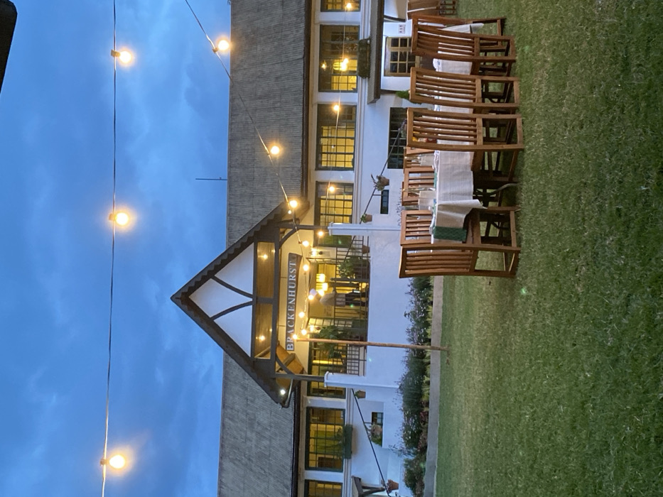 This photograph, taken in Riara Ridge, Kenya, captures an outdoor evening scene featuring a white, two-story building with a distinctive gabled entrance. The building, identified by a sign reading "BRACKENHURST" above its main entrance, has numerous dark-framed windows that reflect the interior lighting. Its architectural style incorporates dark timber framing on the gable, reminiscent of half-timbered designs.
Across the deep blue, twilight sky and adorning the building's facade, multiple strands of illuminated string lights cast a warm glow over the scene. In the foreground and extending to the right, a well-maintained green lawn is visible. Arranged on this lawn are several outdoor dining tables, each set with a white tablecloth, glasses, and what appear to be green placemats or napkins. Matching wooden chairs are placed around these tables, suggesting a setting prepared for an outdoor event or dining experience. A blurry figure is vaguely discernible inside the building near the entrance. Potted plants hang from various points on the building and from a wooden pole on the lawn. The overall impression is one of a serene and inviting ambiance at dusk or early evening.