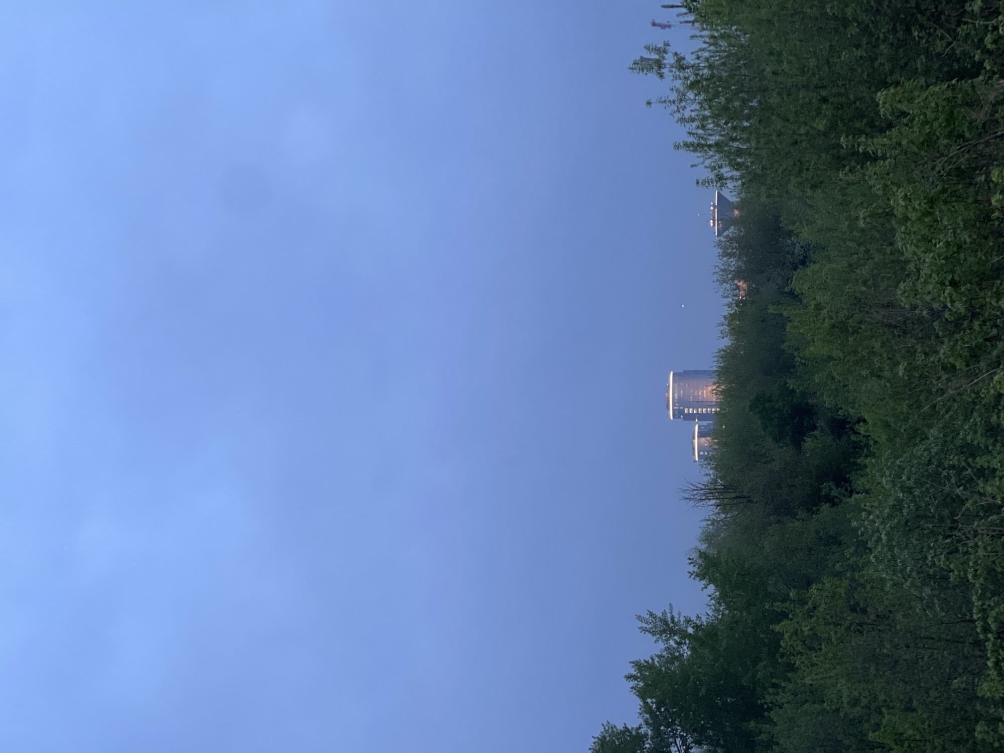 The image presents a view towards the sky with trees lining the right side of the frame. Two tall buildings are visible amongst the trees, with illuminated tops, which place the image in an urban or suburban setting. The sky is a light blue color, suggesting either early morning or late evening. The location is Khimki, Russia, as indicated by the context. There are no visible people or any distinct interactions in the frame.