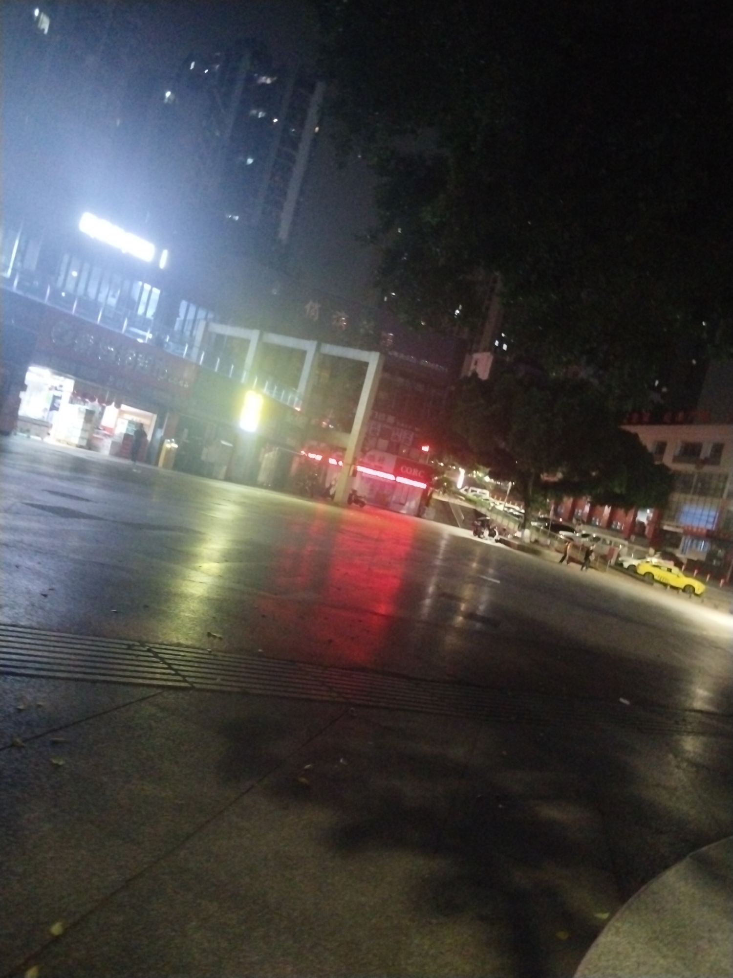 The image captures a street scene in Jiulongpo District, China, at night. The foreground is dominated by a dark, paved area with horizontal grooves. In the middle ground, lit up by bright lights, are several buildings, including a structure with large white arches and a building with a red sign that reads "CORC." Further in the distance, a yellow taxi and other vehicles are visible on a street. People can be seen walking on the sidewalk in the distance near the taxi. Trees are interspersed between the buildings, casting shadows on the scene. A tall building is dimly visible in the background, appearing through the night sky. The lighting appears to be a combination of bright artificial lights and the darkness of night.