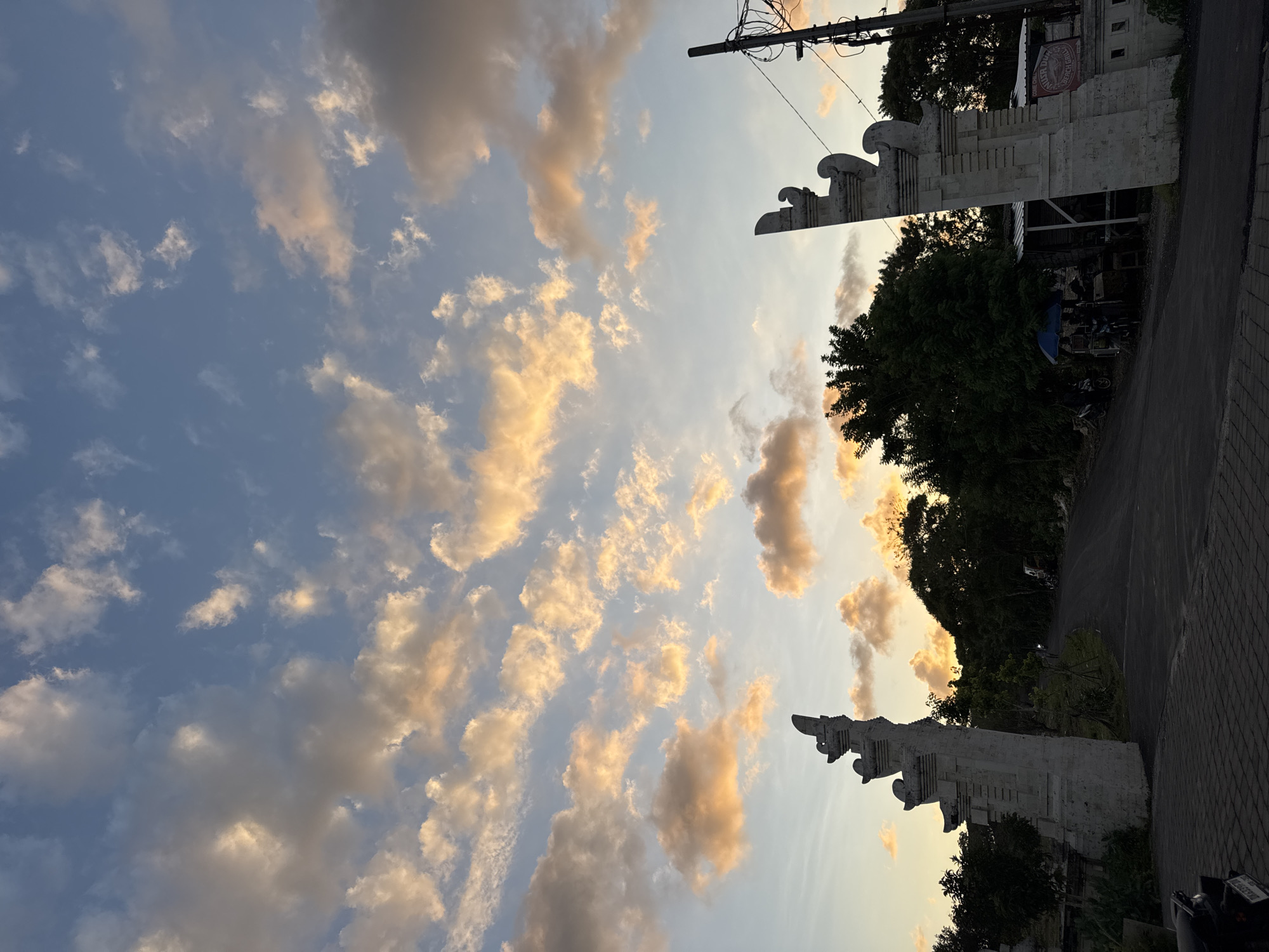 The image shows a view of a street in Kuta Selatan, Indonesia, at dusk. The sky is filled with altocumulus clouds lit by the setting sun, creating a warm, golden hue. On either side of the street stand two ornate, stone structures, possibly Balinese gates, adding to the cultural atmosphere. The road is asphalt, slightly angled, leading to an area with trees and some buildings in the background. There is electrical wiring above.