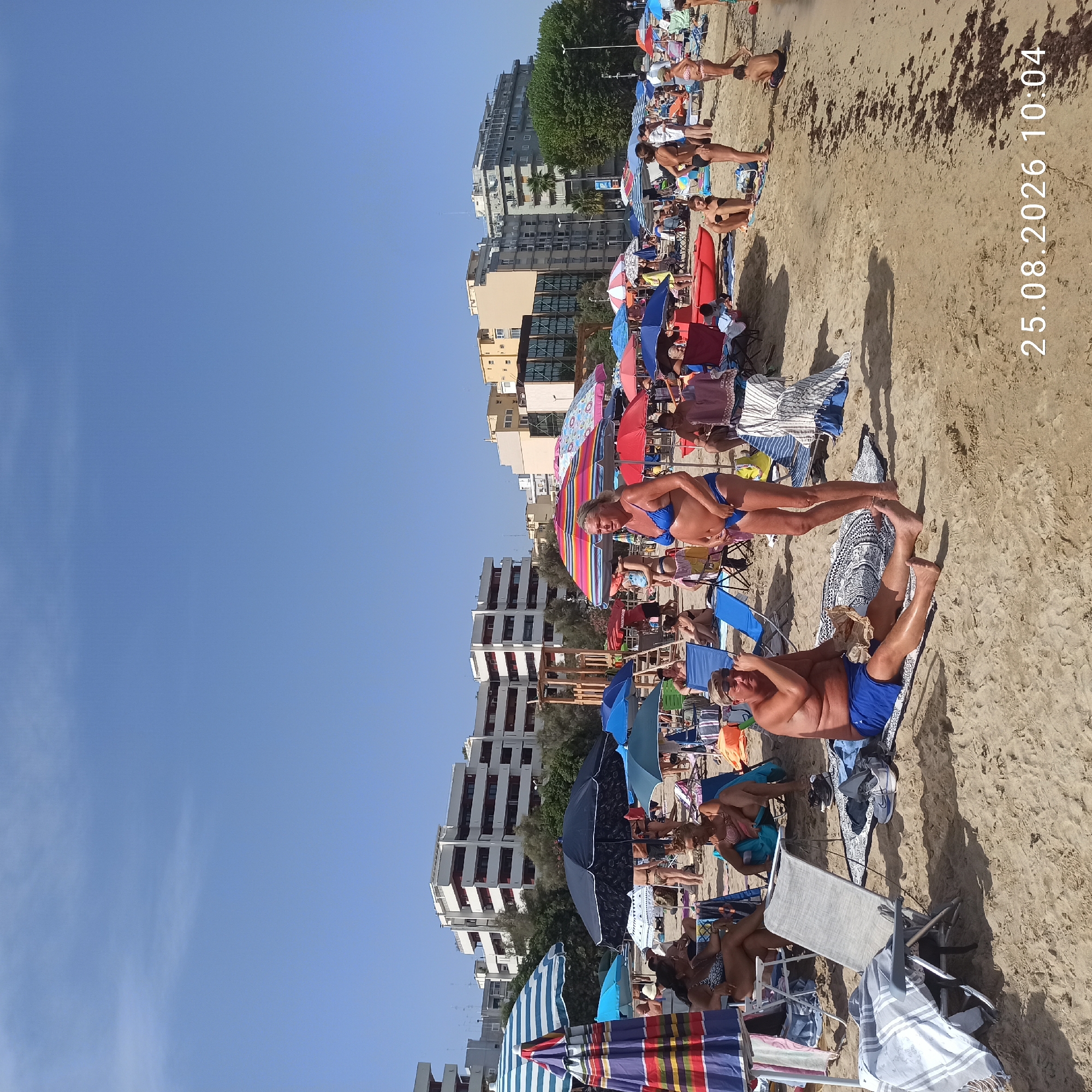 The image depicts a crowded beach scene on a sunny day. In the foreground, a man in blue swim trunks sits on a patterned mat, appearing to be eating something from a wrapper. Beside him, a woman in a blue bikini stands, looking to her right. Several beach chairs and umbrellas are scattered across the sand. In the background, more people are visible relaxing under umbrellas and walking along the beach. Beyond the beach, multi-story buildings are visible, suggesting an urban setting. The sky is clear and blue, with a few wispy clouds. A date and time stamp in the lower right corner reads "25.08.2026 10:04".
