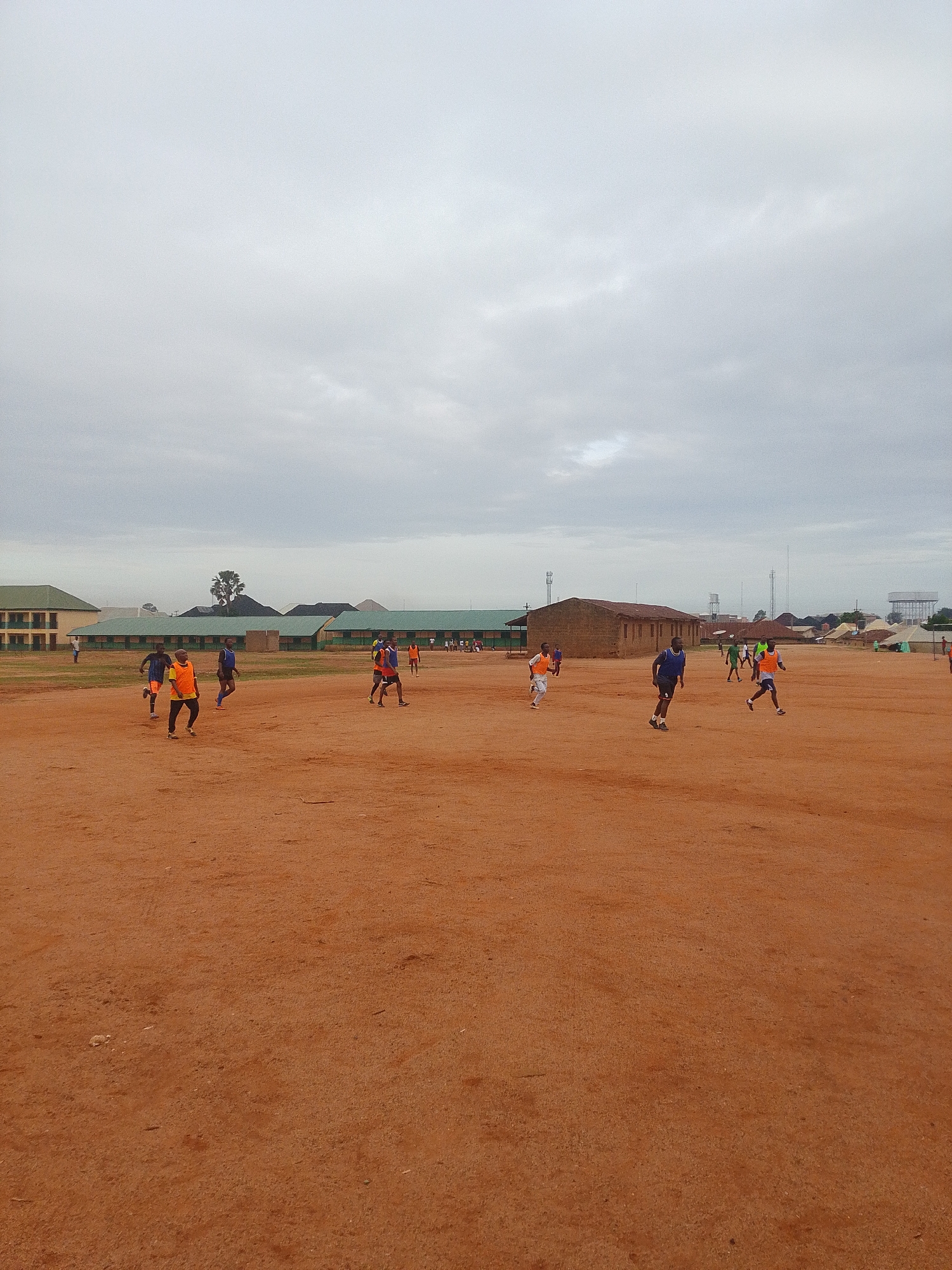 The image depicts a group of individuals playing soccer on a reddish-brown, dirt field. Buildings are visible in the background, including a long, green-roofed structure and a smaller, brown building. The sky is overcast with gray clouds. The location is likely Bukuru, Nigeria. People are running on the field, indicating an active game or practice. Some players wear orange vests over their clothes. In the distance, there are structures that seem to be water towers.