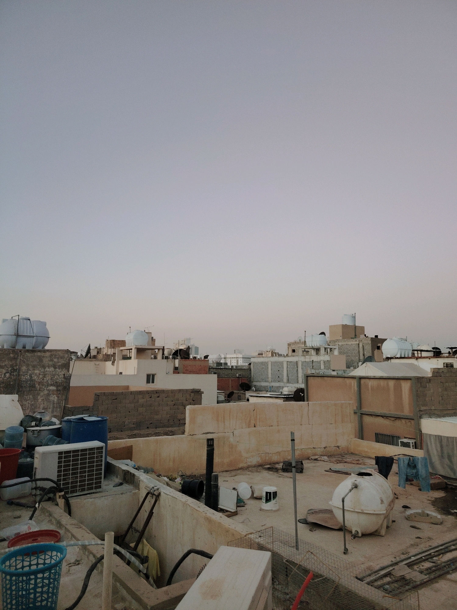 The image depicts a rooftop scene in Riyadh, Saudi Arabia, characterized by a collection of various objects and structures. Dominating the foreground is a concrete rooftop cluttered with a variety of items including large plastic water tanks, a white air conditioning unit, multiple buckets of varying sizes and colors (red, blue, white), and a blue plastic laundry basket. There are also pieces of discarded material, pipes, and what appears to be a small engine or generator. In the mid-ground, more rooftops are visible, featuring brick walls, white and beige painted structures, and numerous water tanks, some white and bulbous, others cylindrical. Satellite dishes are also present on some of the buildings. In the background, the cityscape of Riyadh stretches out with more multi-story buildings. The sky above is a pale, hazy gray, indicating either early morning or late evening light. The overall impression is one of a densely populated urban environment where rooftop spaces are utilized for storage and essential services.