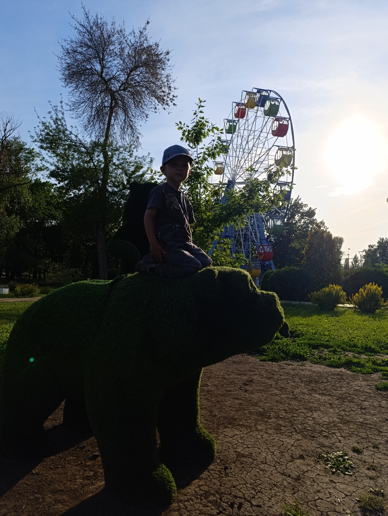 A young boy wearing a blue baseball cap and a dark t-shirt sits on the back of a large, green, topiary bear sculpture. The bear is positioned on a dirt path with cracked soil, surrounded by green grass. In the background, a Ferris wheel with colorful cabins stands against a bright, sunny sky, with trees and bushes flanking the scene. The location is the Central Park of Culture and Recreation in Novokuybyshevsk, Russia.