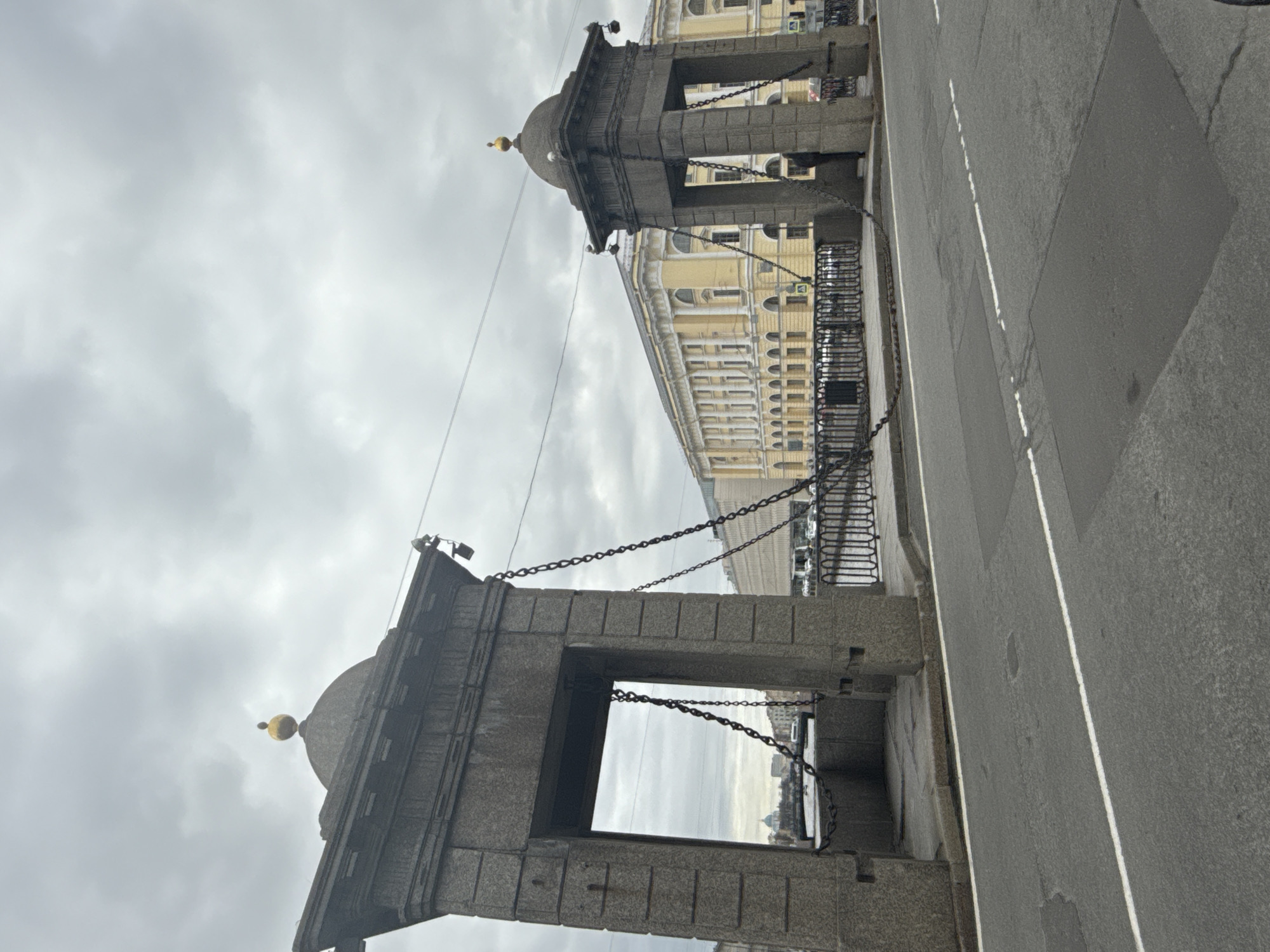 The image captures a static view of a stone bridge in Saint Petersburg, Russia, under an overcast sky. The photograph is oriented vertically, rotated 90 degrees clockwise from its natural horizontal perspective.
In the foreground and mid-ground, two distinctive bridge pylons are prominent. These structures are made of grey stone or granite, featuring a solid, blocky construction. Each pylon has a square base and an arched opening, through which a distant cityscape or river can be glimpsed. The pylons are topped with dark, shallow domes, each crowned with a small, golden spherical finial. Heavy, dark chains are strung between the pylons, extending across the visible span of the bridge.
In the background, a large, multi-story building with a light yellow facade and numerous windows, some arched, is visible. This building exhibits classical architectural elements, contributing to the historical urban setting. To the right, a grey asphalt road surface with faint white lane markings indicates the path across the bridge.
The sky is uniformly grey and cloudy, suggesting an overcast daytime. There are no people, vehicles, or specific activities visible in the scene. The overall atmosphere is quiet and still. No visible text is present within the image.