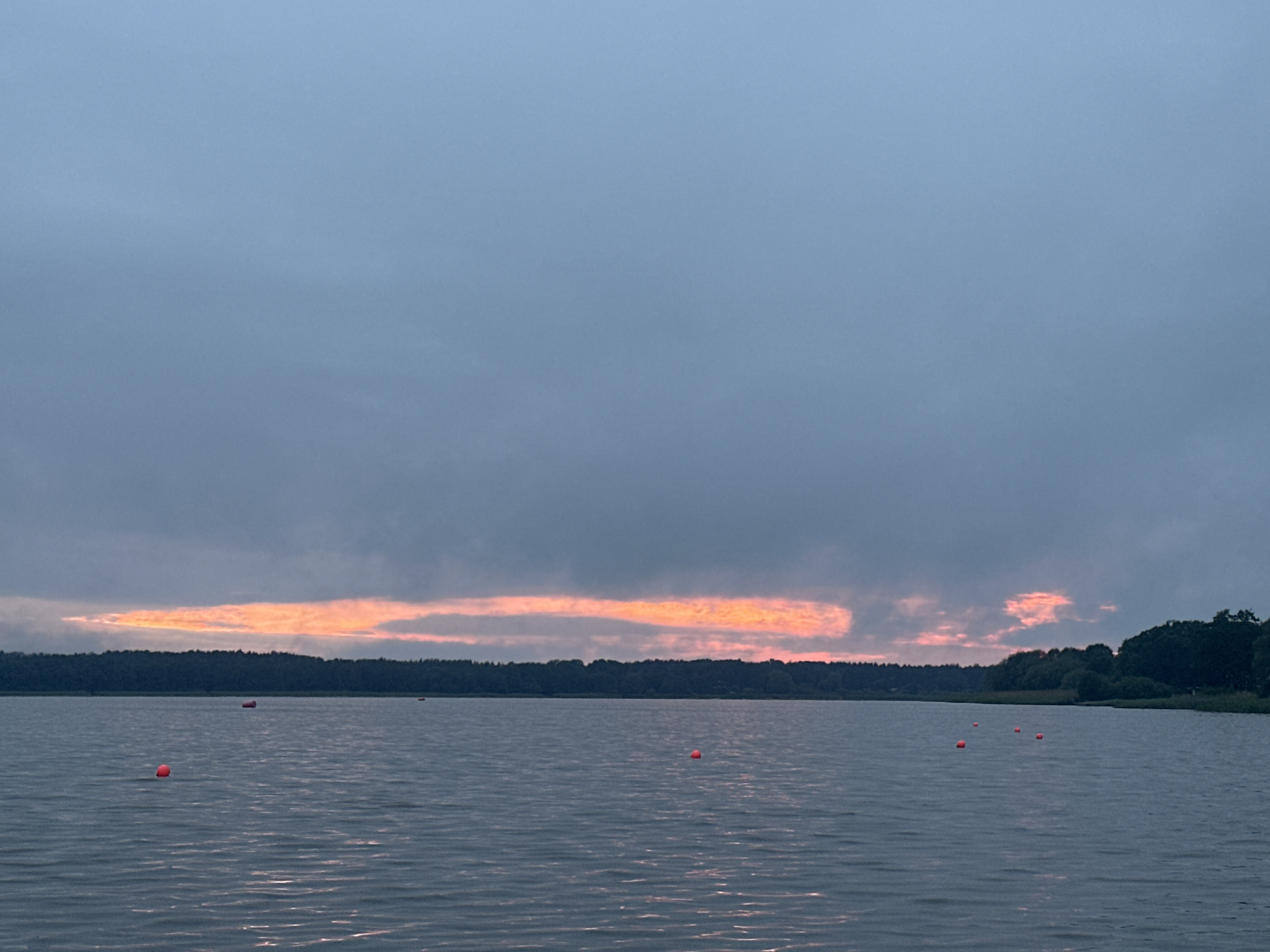 The image depicts a serene lake scene at dusk, with a sliver of orange and pink sunset breaking through the overcast sky. A dark, tree-lined horizon separates the sky from the water. Several small, red buoys are scattered across the lake's surface. The water is calm, with subtle ripples reflecting the muted light. The overall impression is one of tranquility and natural beauty. The location is not specified within the city of Tallinn, Estonia, but rather depicts a general lake environment.