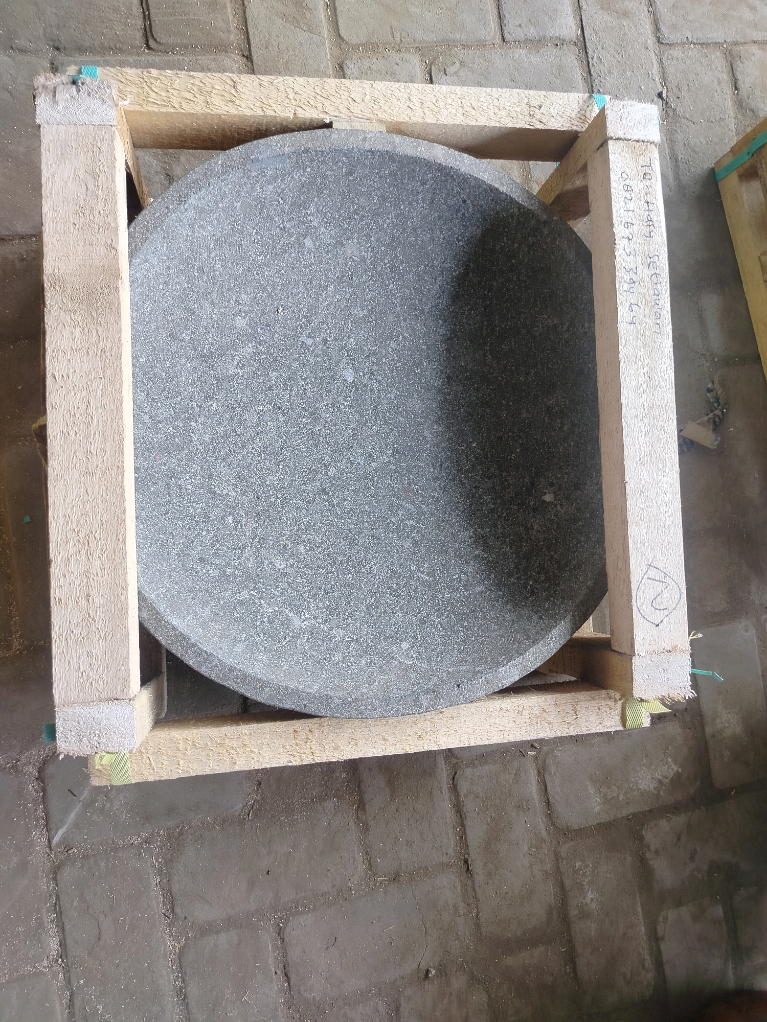 A circular, rough-textured, gray stone object, possibly a millstone, is secured within a wooden crate. The crate is constructed from light-colored wood planks, forming a rectangular frame around the stone. Some of the wood shows rough edges and saw marks. There is some chalk or marker writing on one of the vertical planks of the crate, which appears to be a string of characters and numbers, possibly a label or inventory code, and a circled number "2". The stone object is large and appears to be resting on the bottom planks of the crate, with the sides of the crate providing support. The scene is set on a floor made of rectangular, gray paving stones, which are slightly uneven and appear dusty or dirty, with some scattered wood shavings. The overall impression is one of industrial or construction-related storage.