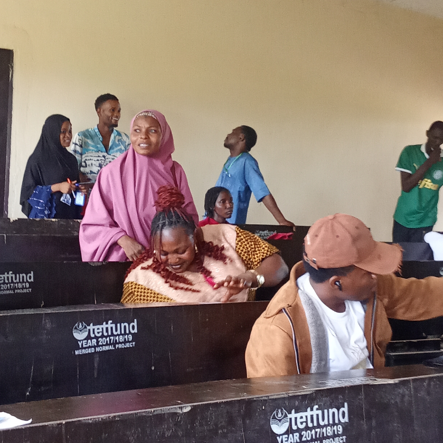 The image depicts an indoor scene, likely a lecture hall or classroom, in Akwanga, Nigeria. Approximately eight individuals, a mix of men and women of various ages, are gathered, some seated and others standing within a tiered seating arrangement. The setting suggests an educational institution or a formal meeting space.
In the foreground, on the right, a man wearing a brown jacket, a white t-shirt, and a brown baseball cap is seen, looking to his left and wearing an earbud. Further back, a woman with braided hair and a patterned top is looking downwards. Centrally, a woman in a pink hijab looks forward with a slight smile. To her left, another woman wearing a black hijab holds papers while looking towards a man in a blue and white patterned shirt, who is smiling. In the background, a man in a blue shirt gazes upwards, while another man in a green sports jersey (possibly a football club shirt) stands on the far right, with his hand near his mouth.
The natural lighting suggests it is daytime. The overall atmosphere appears relaxed and engaged, with several individuals smiling or in conversation.
A prominent detail visible on the dark fronts of the tiered desks is the text "tetfund," repeated multiple times. Below this logo, the phrases "YEAR 2017/18/19" and "MERGED NORMAL PROJECT" are clearly legible. This indicates that the facility was a project funded by the Tertiary Education Trust Fund (TETFund), a Nigerian government intervention agency for tertiary education, during the 2017/2018 or 2018/2019 academic year, reinforcing the educational context within Akwanga, Nigeria.