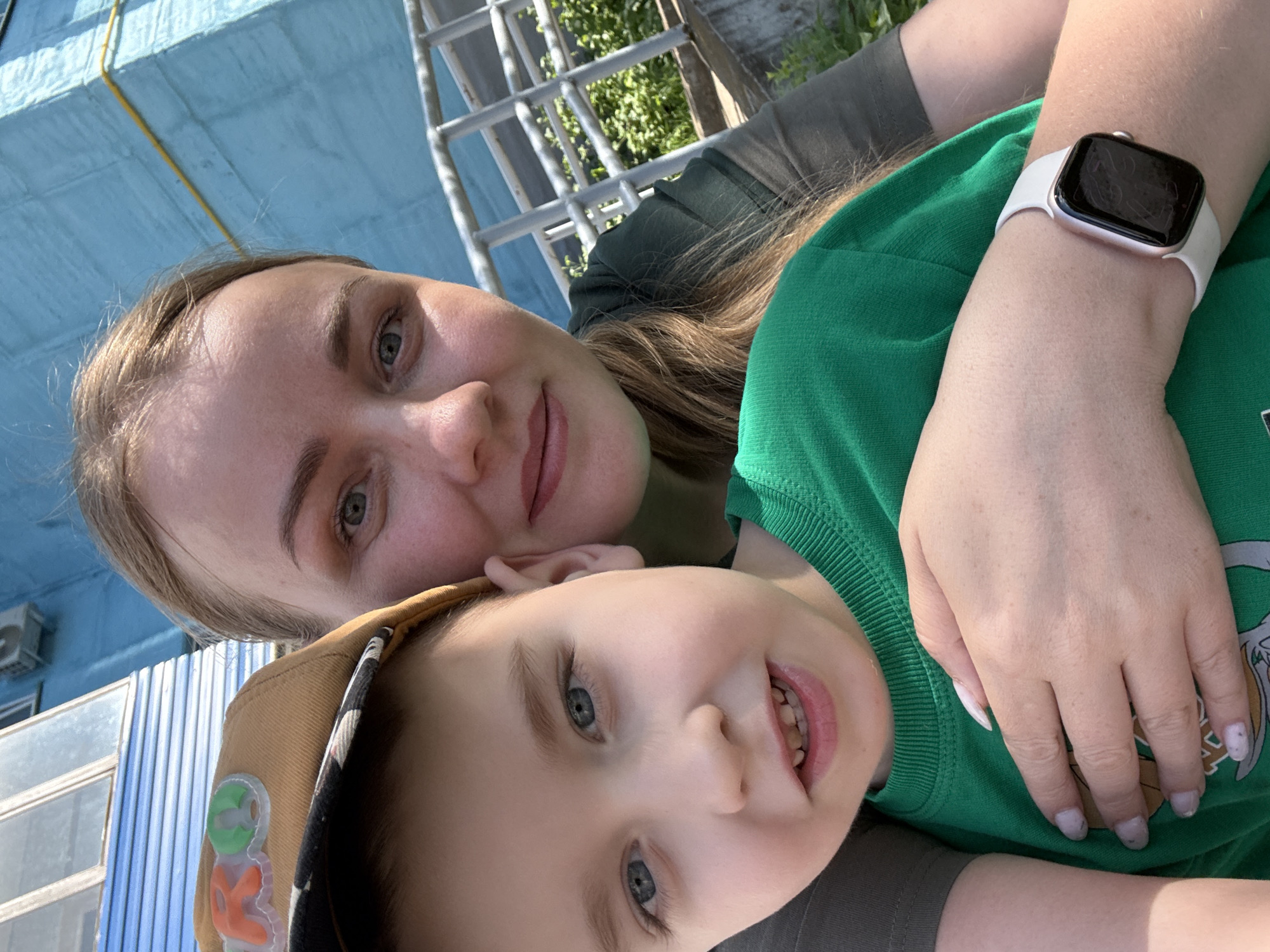 A medium-shot selfie shows a woman and a child, presumably in Chaikovsky, Russia. The woman, with fair skin and blonde hair, is wearing a green t-shirt and has a white smartwatch on her left wrist. The child, with light skin and blonde hair, is wearing a brown baseball cap and a green t-shirt. The child is smiling, revealing their teeth. They appear to be leaning against each other. The background includes a light blue building, a metal structure, and some greenery.