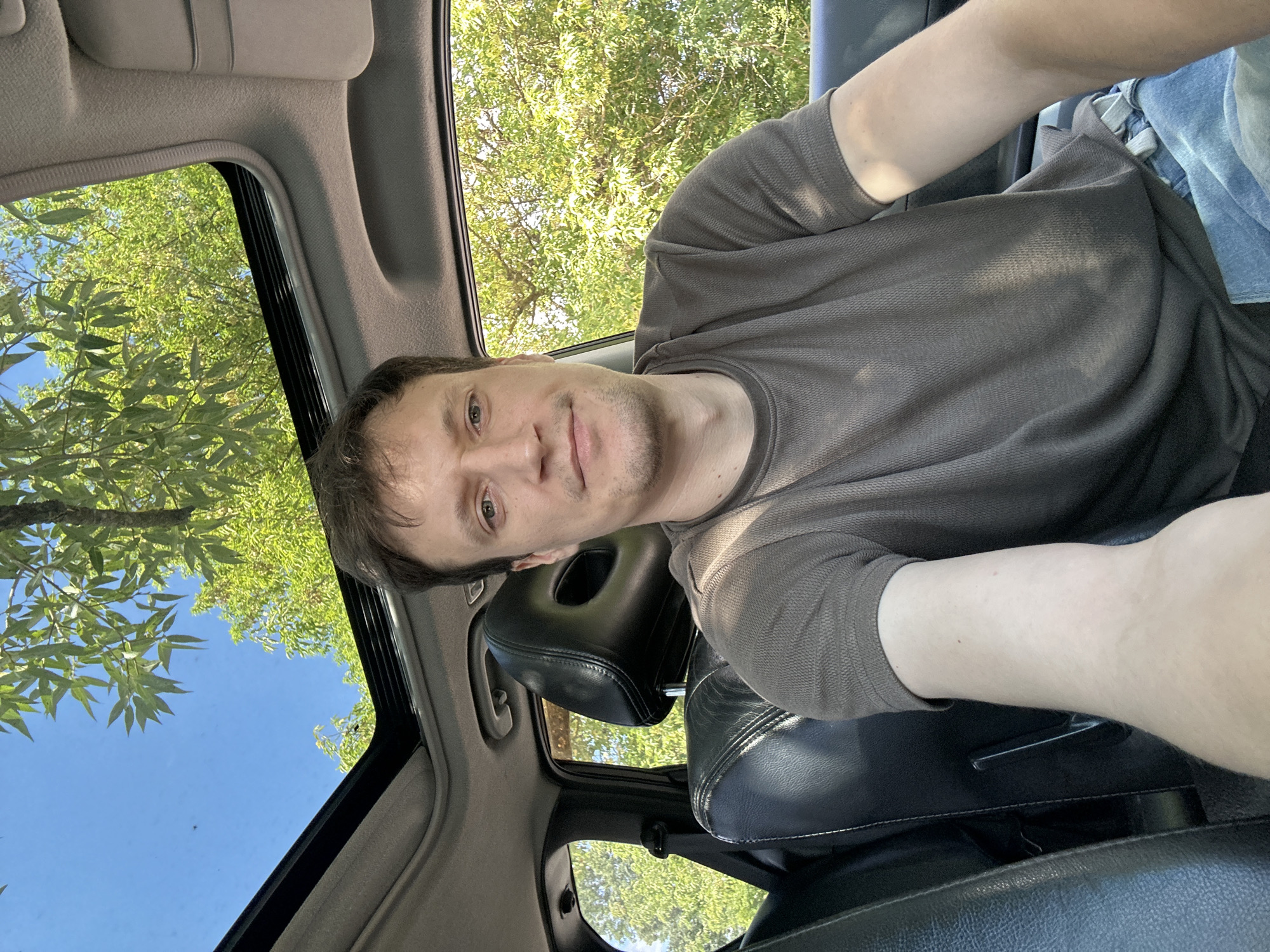 The image shows a man sitting in the driver's seat of a car, with the sunroof open, looking towards the camera. He has short, brown hair, light-colored eyes, and is wearing a dark brown t-shirt and blue jeans. The interior of the car is visible, including black leather seats and the car's headliner. Outside the car, through the open sunroof and side windows, lush green trees and a clear blue sky are visible. The overall setting suggests a sunny day, possibly in a rural or suburban area. There is no indication of the specific location in Makiv, Ukraine, within the image itself.