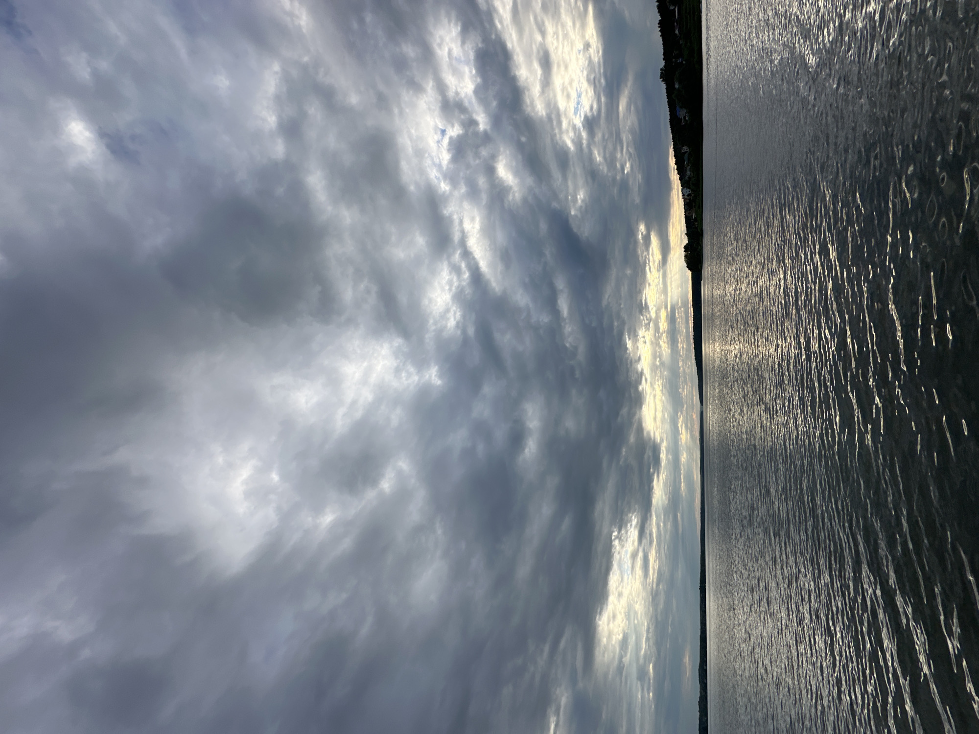 This is a vertical, medium-shot, eye-level photograph of the Votkinsk Reservoir in Votkinsk, Russia, taken on a cloudy day. The sky is filled with dramatic, dark gray clouds that are partially broken, allowing shafts of sunlight to pierce through and illuminate the water. The water's surface is calm with ripples reflecting the light and the sky. In the distance, along the shoreline, there is a line of trees and a cluster of houses, suggesting a populated area. No people or animals are visible in the frame. The overall mood of the image is serene, yet with a hint of dramatic weather.