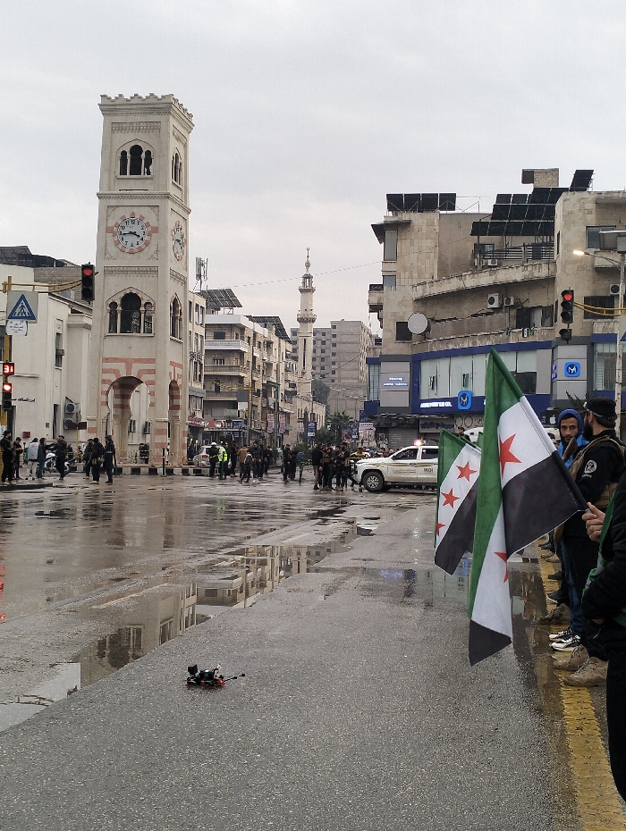 A city scene on a wet day features a clock tower and a mosque minaret in the background. People gather across a road, which is wet with puddles. Flags with red stars are being held in the foreground. A small black and red object lies on the road. Buildings line the street, some with solar panels on the roof. A white police car is visible amidst the crowd.