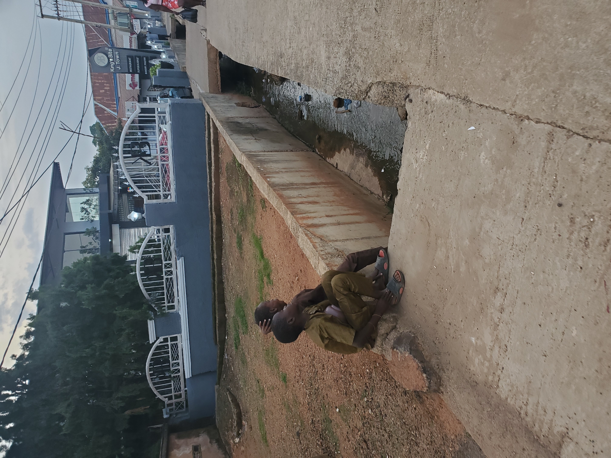 Two young boys, appearing to be of primary school age, are seated on a concrete ledge overlooking a shallow, debris-filled drainage channel. They are both wearing simple, loose-fitting clothing. The boy in the foreground is dressed in a tan or olive green outfit and is looking to his right, with his left hand resting on his knee. The second boy, behind and slightly to the right of the first, is wearing a darker brown outfit and has his left arm raised, with his hand touching the back of his head. They are positioned on the edge of a concrete pathway that runs alongside the channel.
In the background, across the drainage channel, is a modern building with large glass windows and a dark roof. A grey painted wall with white wrought-iron style fencing runs along the property line. Beyond this building, a street is visible with other commercial establishments and overhead electrical wires. One sign clearly reads "King's Bite J. Town" and lists "RESTAURANT," "SUPERMARKET," and "BAKERY/PASTRY." The overall environment suggests an urban or semi-urban setting. The sky is overcast, indicating a cloudy day. The scene appears to be located in Jos, Nigeria, as suggested by the presence of a business named "J. Town," which is a common abbreviation for Jos.
