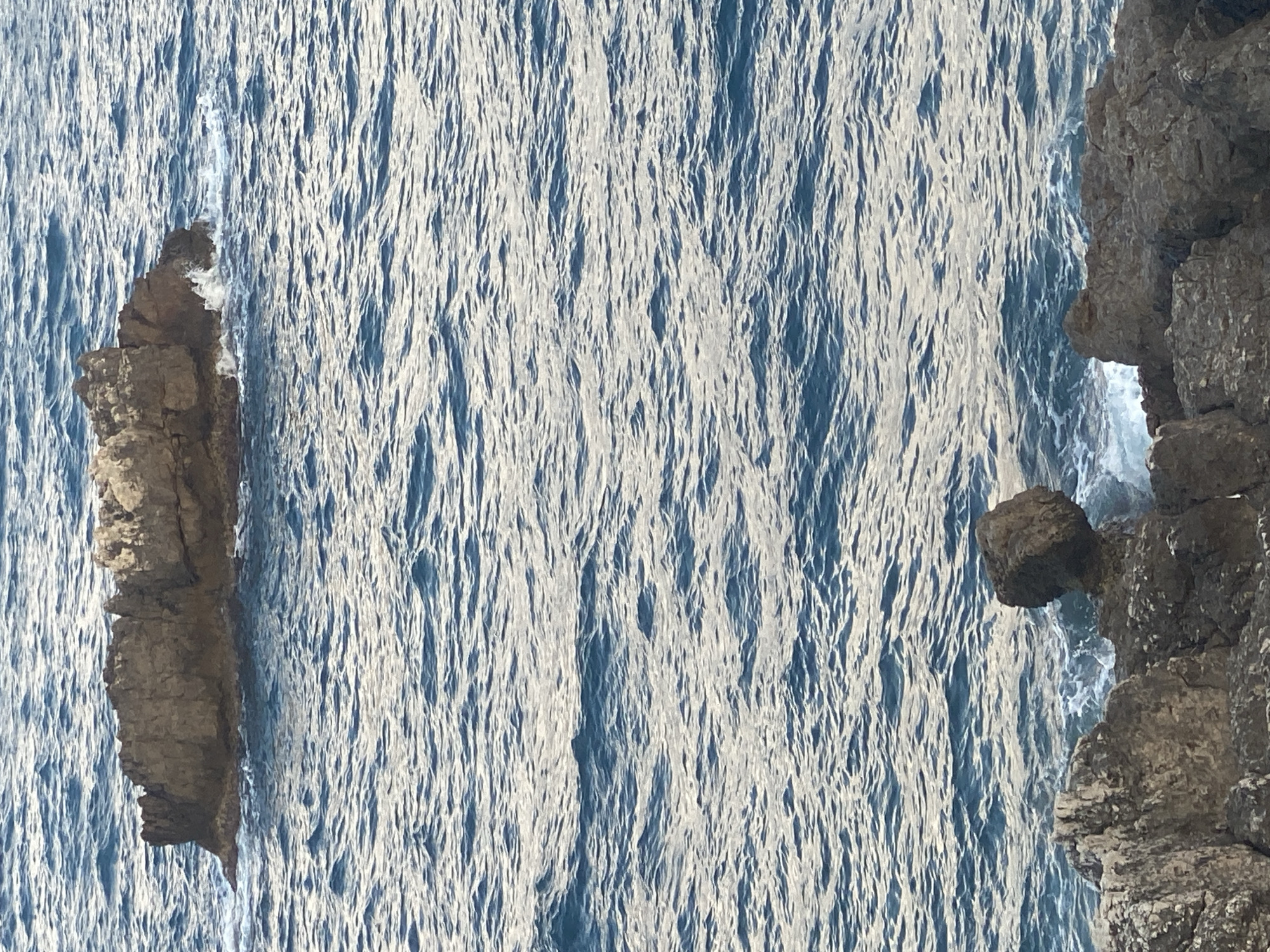 A medium shot captures a rocky outcrop in the Mediterranean Sea off the coast of Antibes, France. The water is a deep blue, with gentle waves lapping against the rocks and creating frothy white accents. The rocks themselves are dark and weathered, with some sharp edges and craggy surfaces. The scene is serene and natural, with no human presence or artificial elements visible. The time of day appears to be late afternoon or early evening, as indicated by the soft, diffused light.