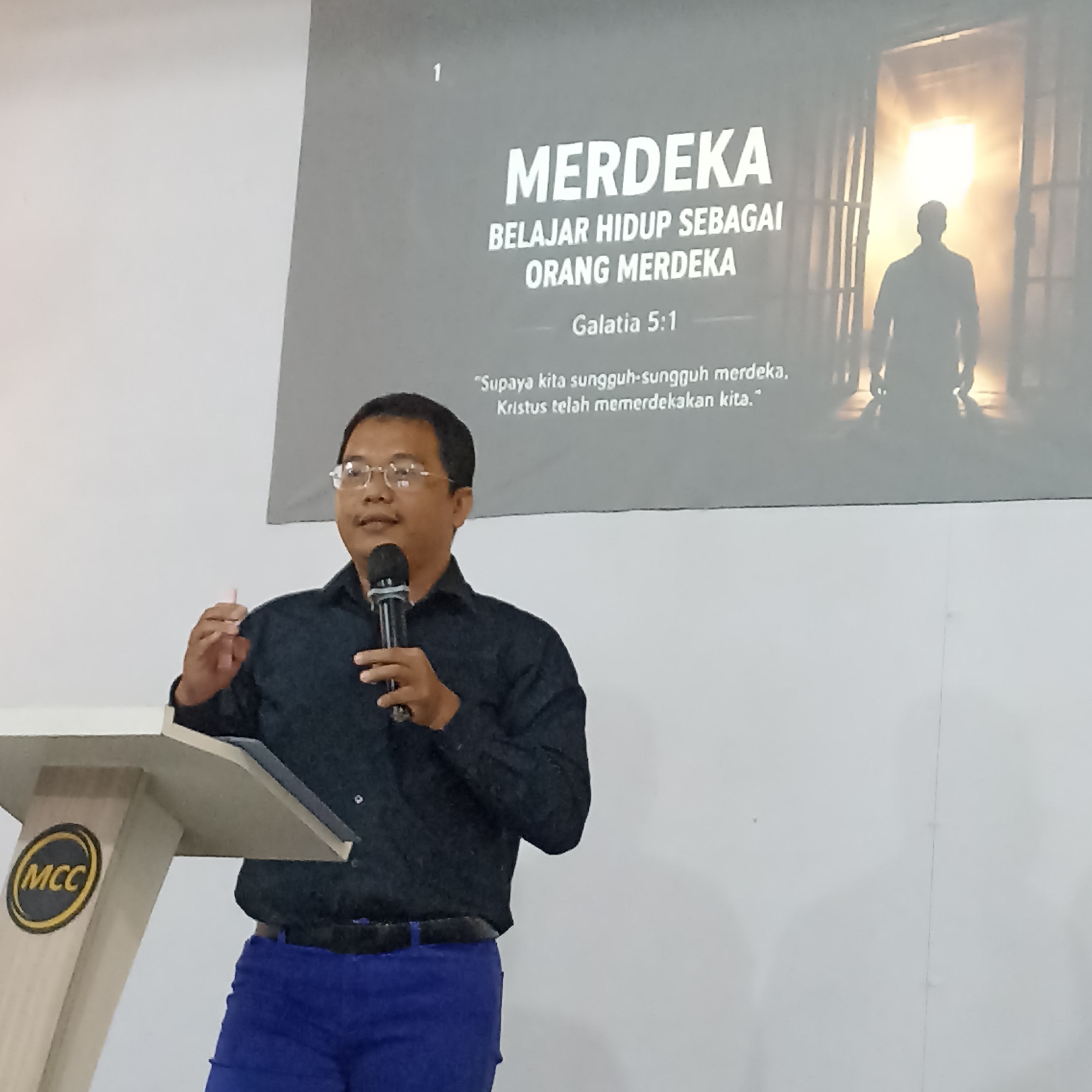 The image shows a man wearing a black shirt and blue pants, standing behind a podium with the "MCC" logo on it. He is holding a microphone and appears to be speaking. Behind him, a projection screen displays text and an image. The text on the screen reads "MERDEKA BELAJAR HIDUP SEBAGAI ORANG MERDEKA" followed by "Galatia 5:1" and a quote in Indonesian. The image on the screen depicts a silhouette of a person walking towards an open doorway with bright light emanating from it. The overall setting appears to be an indoor presentation or event. The MCC logo suggests the location is an MCC (Majelis Christus Community) institution, which has branches in Magelang, Indonesia.