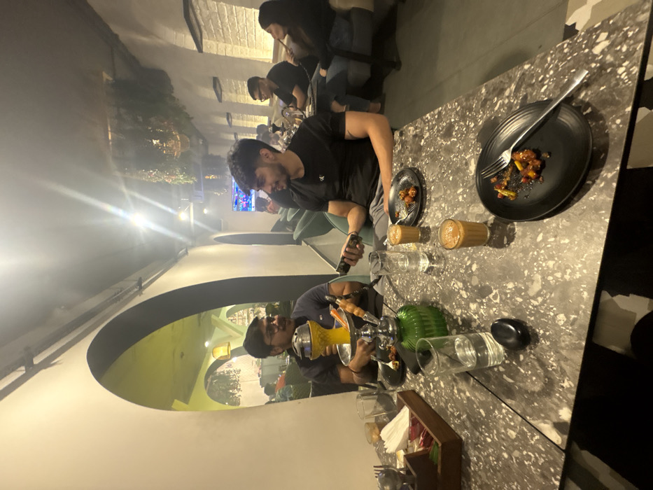 A group of people sit at a table. One man holds a phone, near food and drinks. Another has a hookah pipe. Black plates with food remnants and a fork sit on the patterned table. Glasses of water, small teacups, and hookah components are also present. The setting appears to be an outdoor patio or restaurant, possibly with television screens in the background. Arched architecture is visible in the background.
