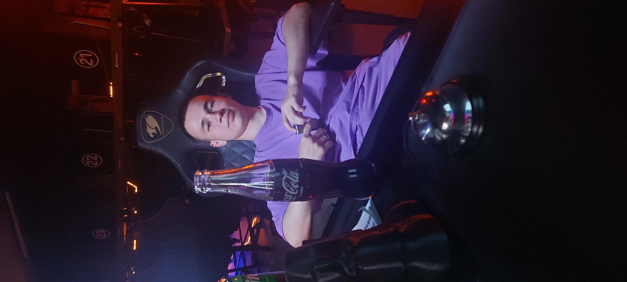 A male wearing a purple shirt is reclining in a black Cougar brand gaming chair. A glass Coca-Cola bottle is positioned near his lap, and a silver bell sits on a dark surface to his right. Behind the individual, numbers "21", "22", and "23" are visible, possibly indicating seating positions within an entertainment venue. The setting appears to be a gaming or internet cafe in Tashkent, Uzbekistan, given the gaming chair, numbered seating, and overall dimly lit atmosphere.