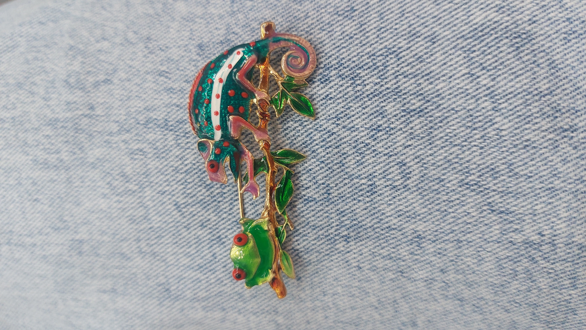 A close-up shot depicts a chameleon and frog brooch pinned to blue denim fabric. The brooch features a green and pink chameleon with red spots clinging to a gold-colored branch. A green frog with red eyes sits below the chameleon on the same branch. The denim fabric is visible in the background. There is no information about the location in Novosibirsk, Russia, in the image.