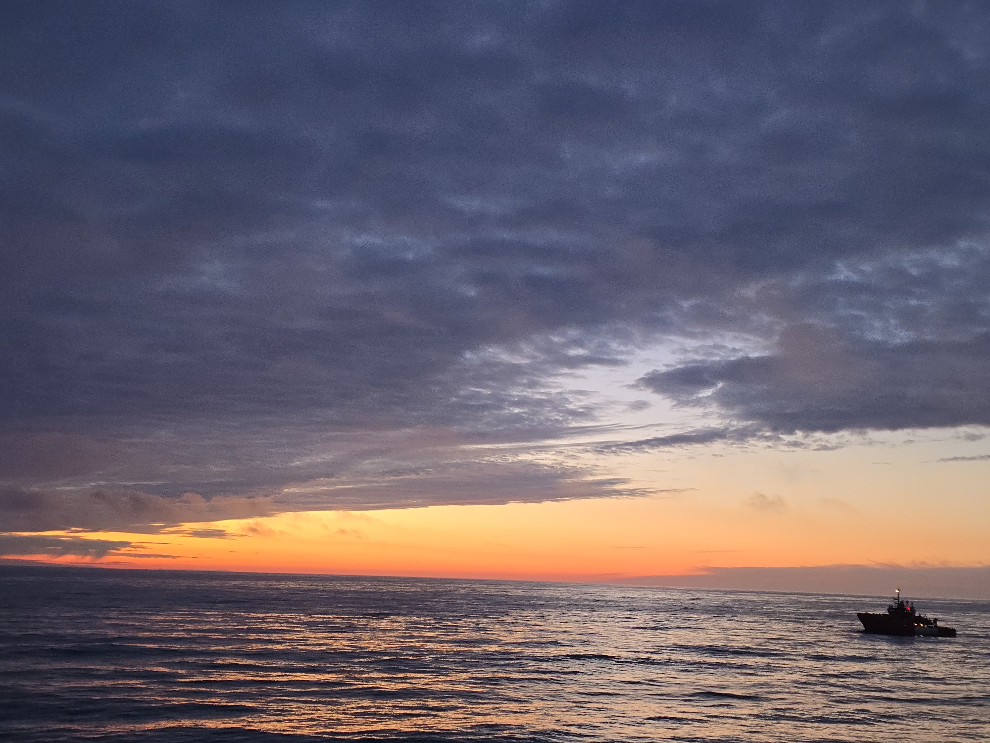 The image shows a body of water with a boat, with a sunset or sunrise. The sky is partially covered by grey and purple clouds. A thick layer of orange-yellow light illuminates the horizon. The boat is located in the right of the frame. The location is not specified.