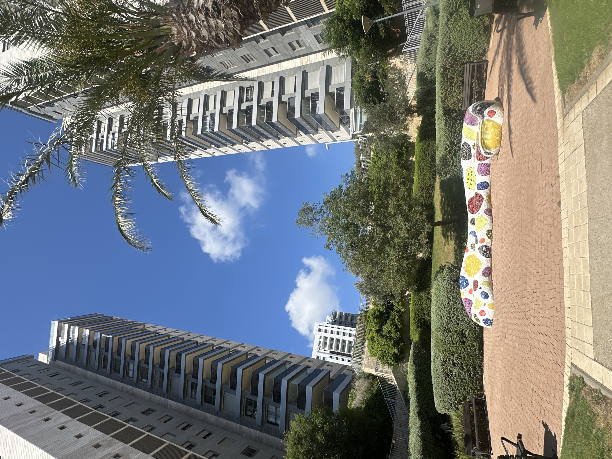 This is a media file captured in Haifa, Israel. The scene depicts a residential area with modern high-rise buildings against a clear blue sky with scattered white clouds. A palm tree is visible in the foreground, with its fronds casting shadows. The buildings are characterized by their sleek, contemporary design, featuring balconies with angled sunshades and many windows.
In the mid-ground, there are well-maintained green hedges and trees, suggesting a park-like setting within the urban environment. A paved walkway, made of reddish-brown bricks, runs alongside the greenery.
A prominent object in the foreground is a large, colorful sculpture. It appears to be shaped like an elongated, curved object, possibly a stylized animal or abstract form. The sculpture is covered in a mosaic of brightly colored tiles in various shapes, including yellow, red, blue, purple, and white. One section of the sculpture, resembling a head, has black and red tile details.
A park bench is situated near the sculpture, and a black trash receptacle is visible further down the path. The presence of the sculpture, benches, and manicured landscaping indicates a public space designed for recreation or aesthetic enjoyment. The lighting suggests it is daytime, likely late morning or early afternoon, given the shadows. No people are visible in the frame.