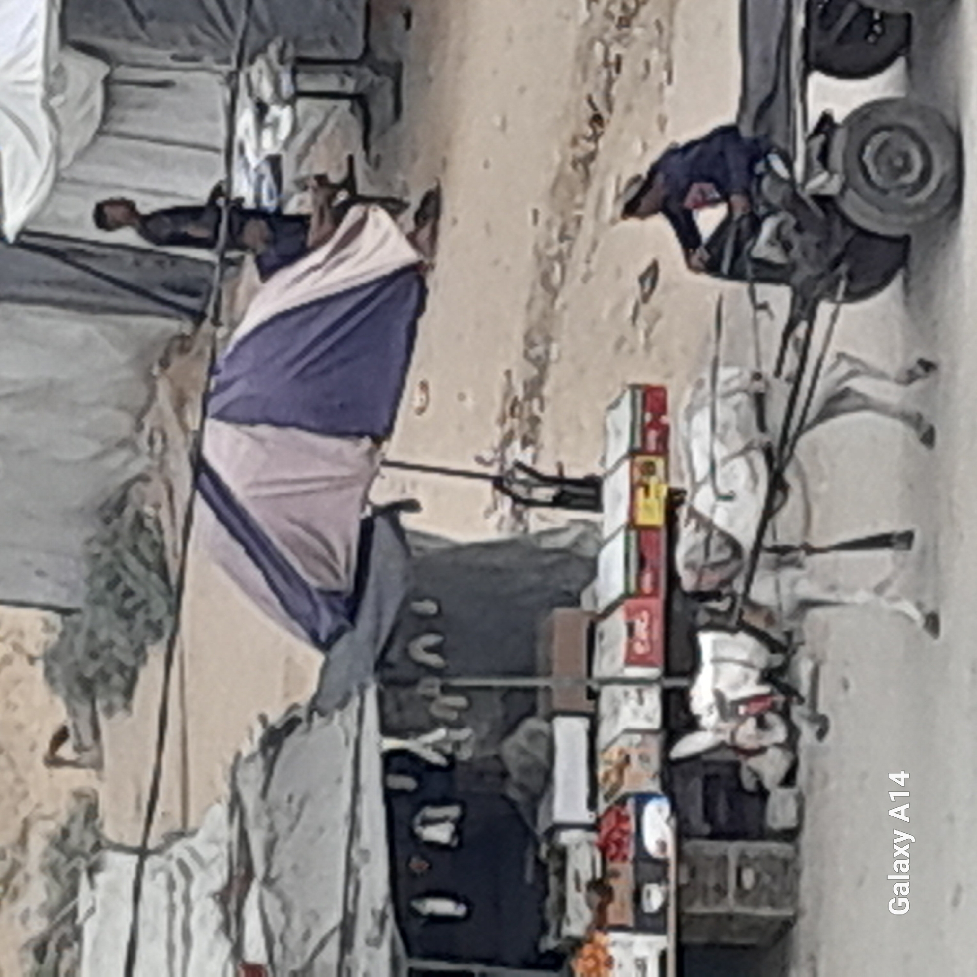A donkey pulls a cart laden with sacks through a sandy street in Khan Yunis, Palestinian Territories. The scene depicts a market setting with stalls and tents, suggesting a bustling commercial area. A man is seated on the cart, guiding the donkey. The donkey is adorned with a red and white halter, and its back is covered with a patterned blanket. The cart itself is dark and functional, with large wheels. In the background, a man in a dark shirt stands near a stall displaying various items, possibly goods for sale. The overall environment appears arid and dusty, characteristic of a region with limited rainfall. The presence of tents and market stalls indicates a temporary or semi-permanent settlement, possibly associated with informal trade or aid distribution. The "Galaxy A14" watermark is visible in the lower left corner, indicating the device used to capture the image.