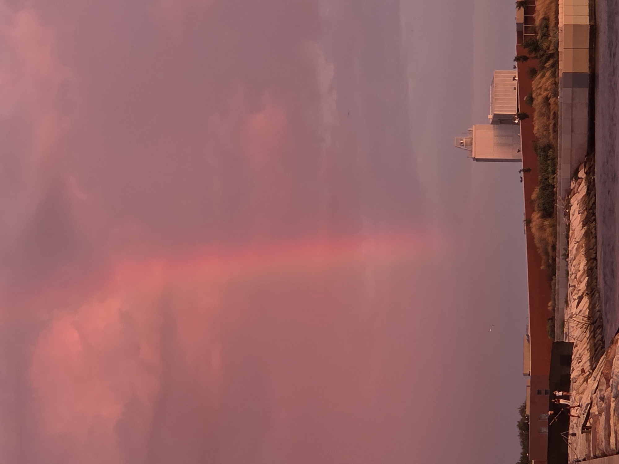 A photograph taken from the waterfront in Chioggia, Italy, captures a dramatic sunset with vibrant pink and purple clouds. A faint rainbow arcs through the sky. In the foreground, a stone pier juts out into the water, with two figures standing on it, seemingly fishing. Behind them, a low brick wall runs along the embankment, topped with dry, golden grass. Further in the background, a modern white building with a tower stands against the dusky sky. The water in the foreground is dark and calm, reflecting the colors of the sky.