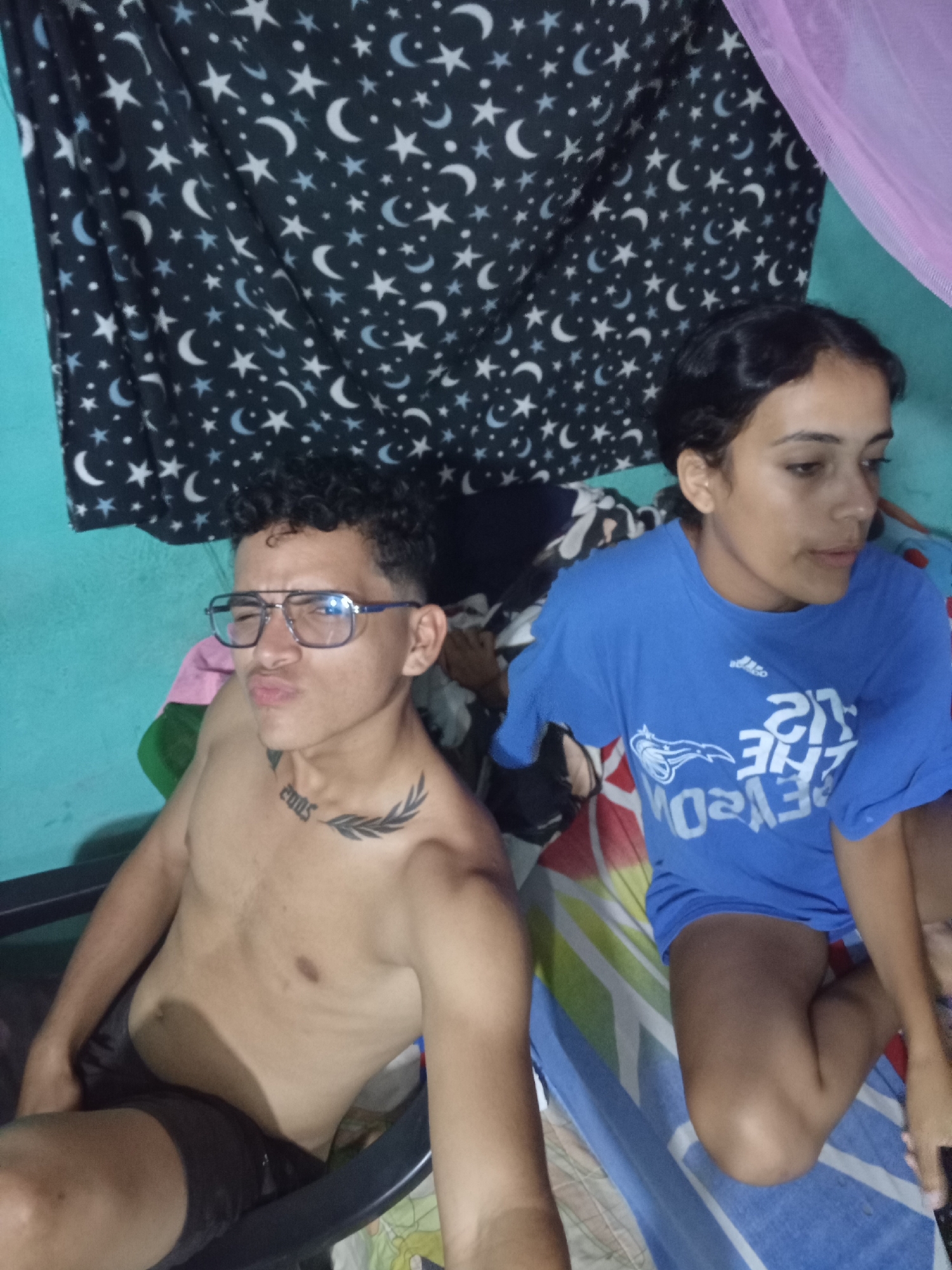 Two individuals are visible in a room. The person on the left is a shirtless young man wearing glasses, sitting in a black chair. He has a tattoo on his chest. The person on the right is a young woman wearing a blue t-shirt. She is sitting on a bed with a striped blanket. The background includes a wall painted in teal and a curtain with a star and moon pattern. Part of a pink fabric, possibly a mosquito net, is visible in the upper right corner. Based on the prompt the location is Altagracia, Venezuela.