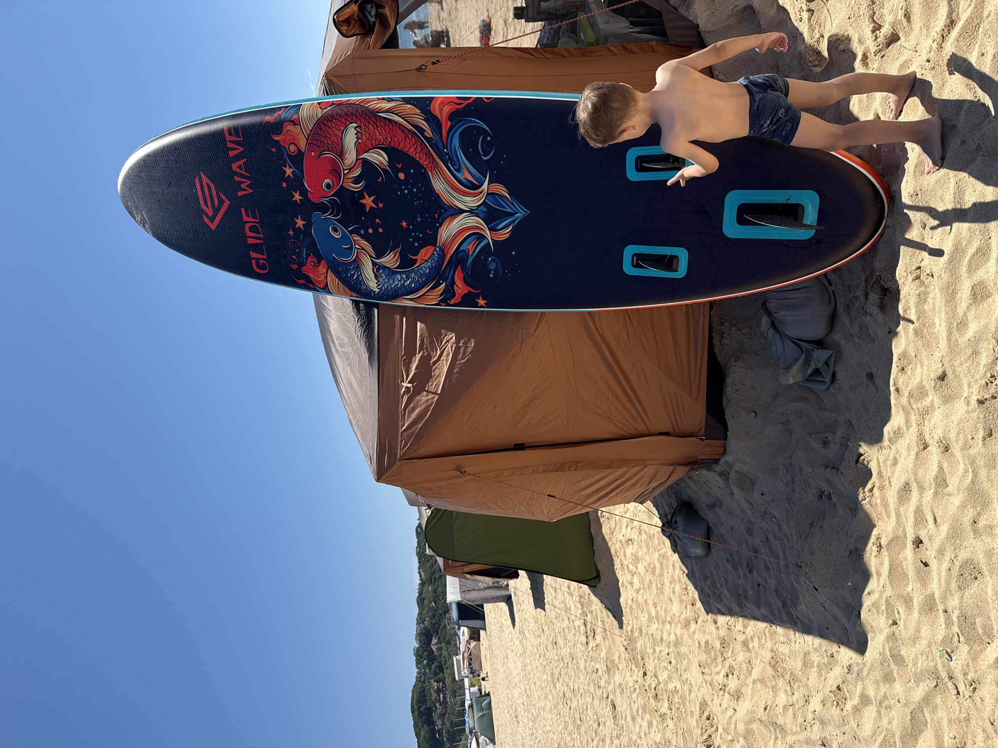 The image depicts a sandy beach scene under a clear blue sky. In the foreground, a young boy, shirtless and wearing dark patterned swim trunks, stands with his back to the camera, facing a large, dark blue stand-up paddleboard decorated with an intricate design of two koi fish, flames, and stars. The paddleboard has two handles with turquoise accents and fins visible at the bottom. Behind the paddleboard and the boy is a large, tan-colored tent, partially obscuring other tents lined up along the beach in the background. Some of these tents are green and grey. The sand is light brown and appears somewhat disturbed, with shadows cast by the tent and paddleboard. In the distance, the sea is visible, with some boats and people along the shoreline. The overall environment suggests a camping or beach outing. The location is not specified as Anna, Russia.