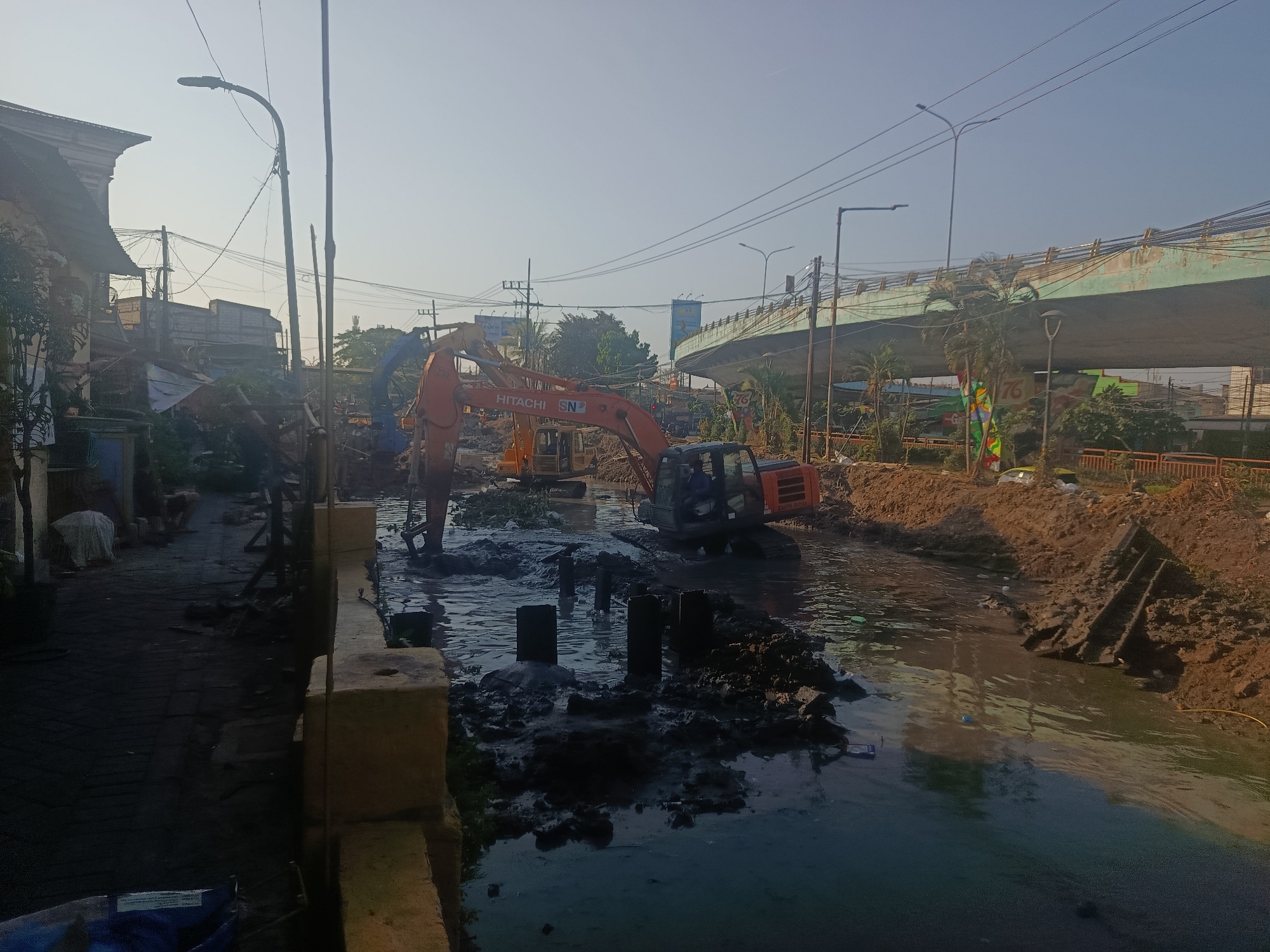 In Surabaya, Indonesia, construction work is underway on a water channel, possibly for flood control or infrastructure development. The scene is dominated by excavators, with one prominent orange Hitachi excavator actively digging in the muddy water. Several dark, rectangular structures, possibly pilings, protrude from the murky water, indicating ongoing foundation work.
To the left of the water channel, a paved walkway runs alongside a building with an ornate facade, suggesting an older architectural style. Debris and construction materials are scattered along the walkway and behind the building, hinting at demolition or renovation. Power lines crisscross the sky, supported by utility poles, and a streetlamp stands to the left.
In the background, an elevated road structure, likely an overpass or bridge, spans across the scene. The sky is clear and bright, indicating daylight. The overall atmosphere is one of active construction and urban development in a dynamic cityscape.