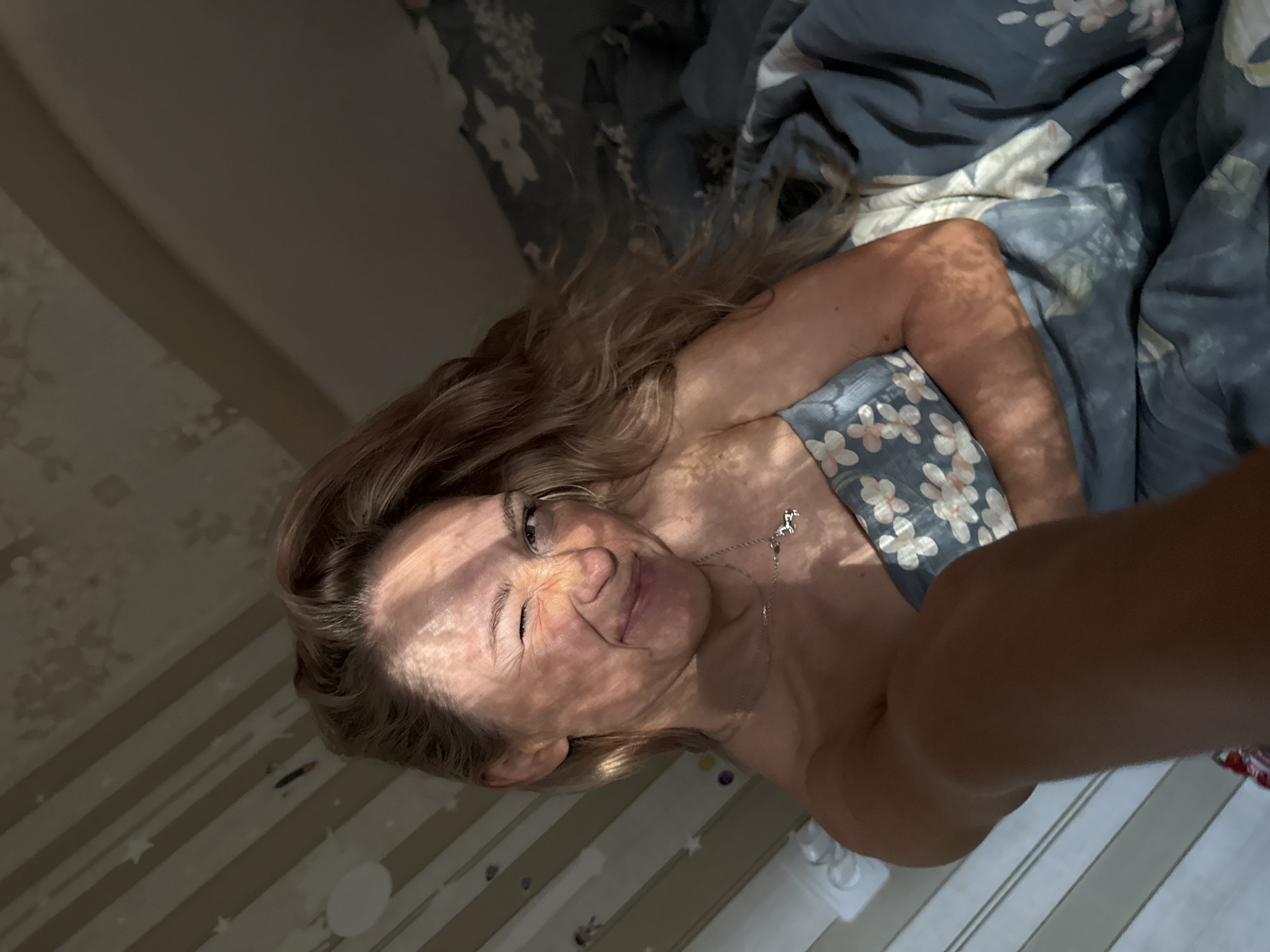 A woman is lying in a bed in what appears to be an indoor location, based on the interior environment. The bed has floral-patterned sheets. The woman has long, wavy blonde hair, and her face is partially obscured by shadows, but she appears to be smiling and winking. She is wearing a shirt or pajamas with a light blue background and floral patterns. A silver necklace with a horse pendant is visible around her neck. The room also shows a striped pattern on the wall. There is no indication of a specific location in the city of Chaikovsky, Russia.