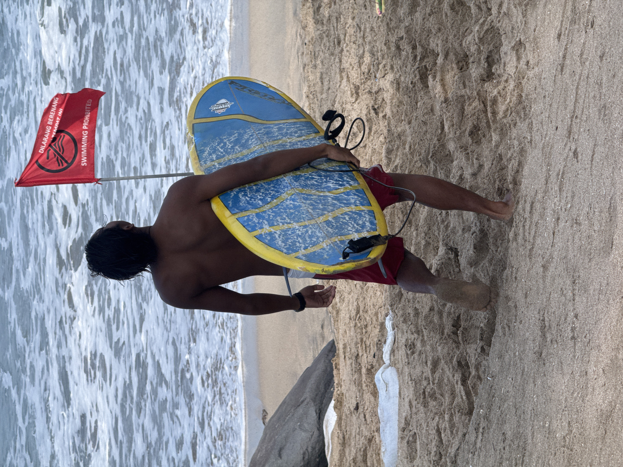 A man with dark hair, facing away from the camera, walks on a sandy beach. He is carrying a blue and yellow boogie board under his left arm. He wears red shorts. Behind him, a red flag with white text and a black symbol is planted in the sand, indicating "SWIMMING PROHIBITED." The ocean with white-capped waves is visible in the background. A large, dark rock is situated to the left of the man. The sand is textured with footprints and some debris. The overall scene depicts a beach with a warning about swimming.